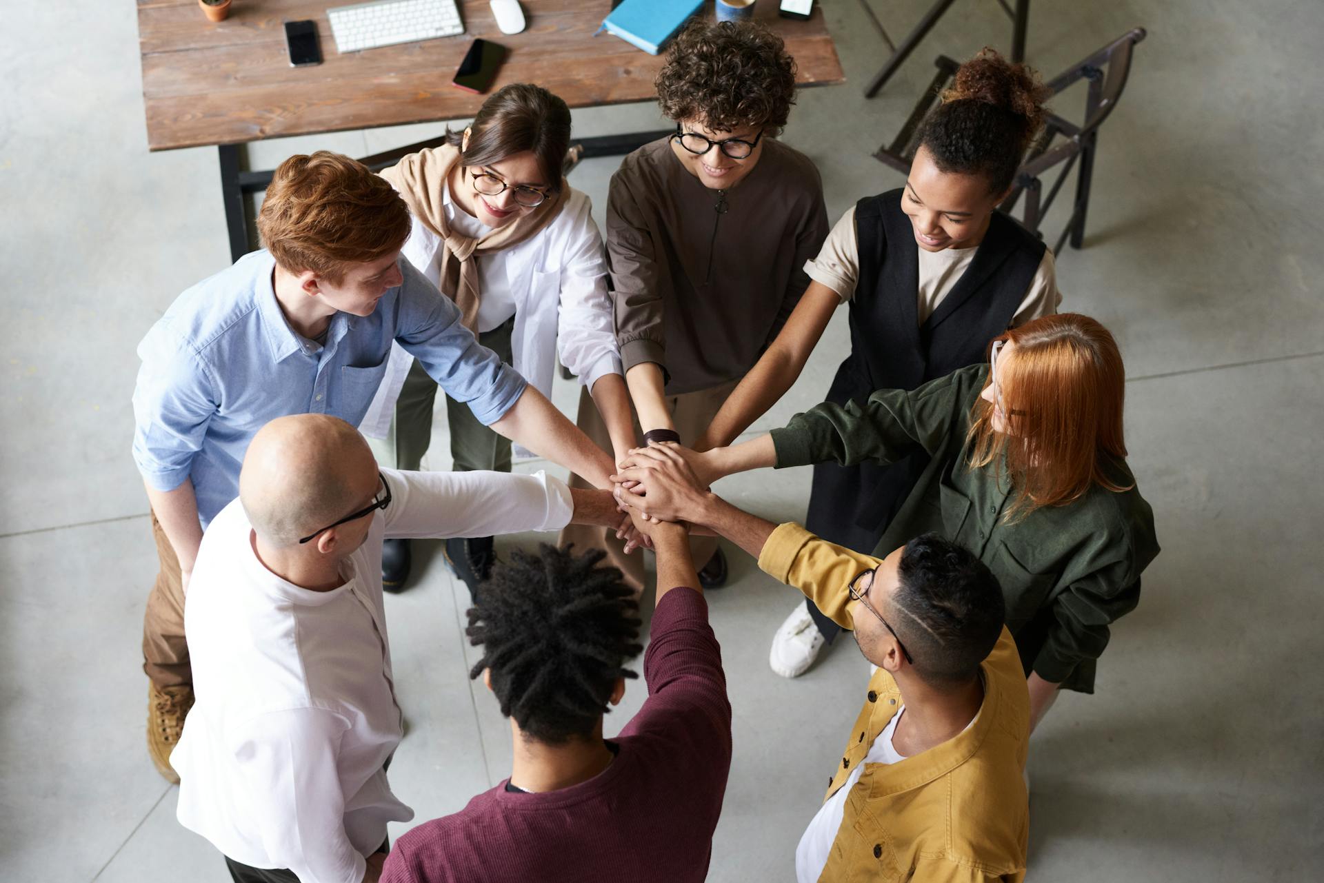 Vogelperspektive auf eine diverse Gruppe von acht Personen, die ihre Hände in der Mitte eines Kreises zusammenlegen – eine Geste des Teamgeists und der Zusammenarbeit. Das Team steht in einem modernen Büroraum mit hellem Boden, im Hintergrund ein Holztisch mit Computern, Notizbüchern und Smartphones. Die Teammitglieder tragen lässige Business-Kleidung in verschiedenen Farben.