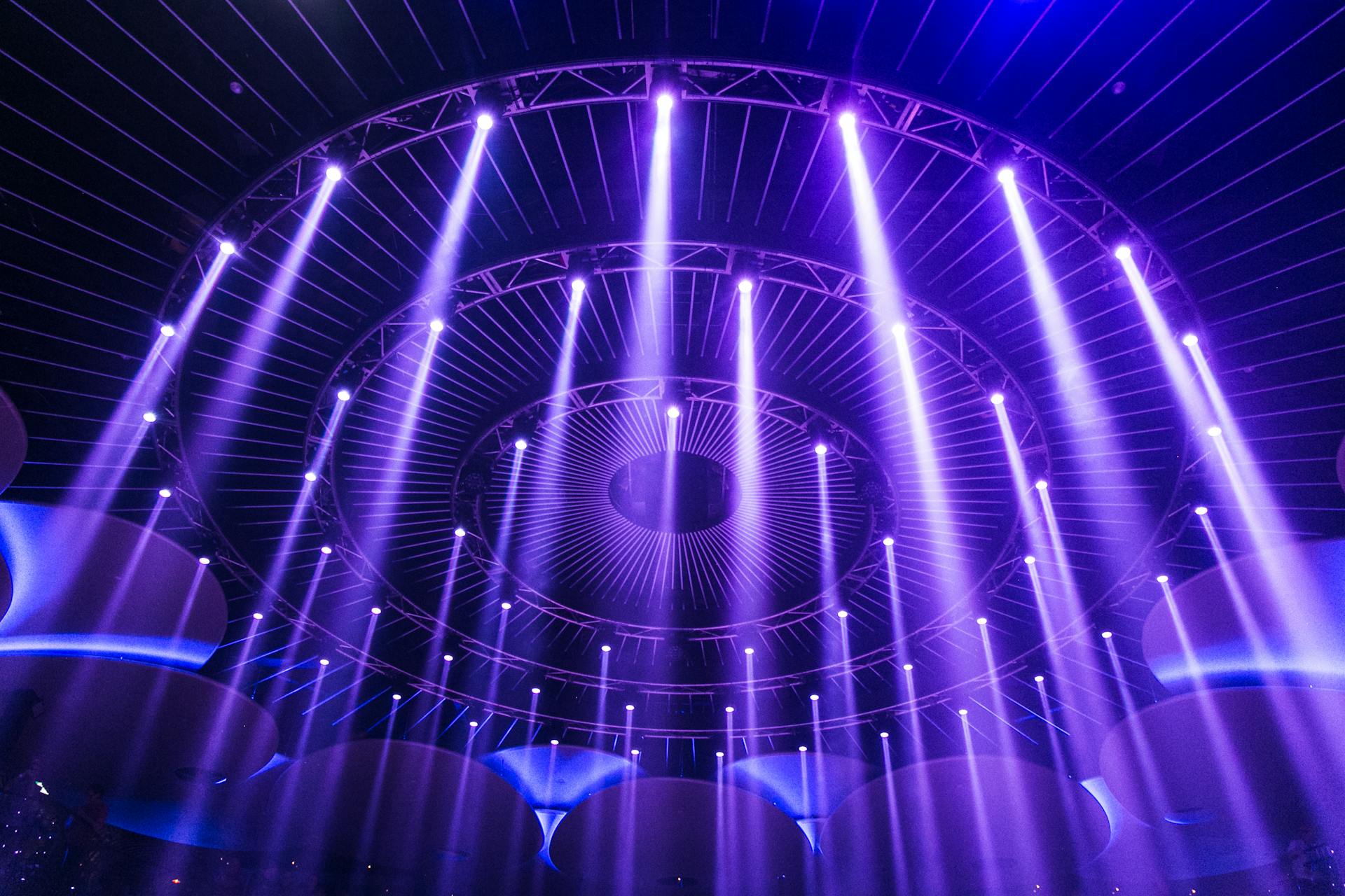 Spektakuläre Bühnenbeleuchtung mit konzentrischen kreisförmigen Lichtinstallationen in intensivem Violett und Blau. Zahlreiche vertikale Lichtstrahlen durchdringen den Raum von einer mehrschichtigen, kreisförmigen Bühnenkonstruktion aus, die an ein technisches Auge oder einen Tunnel erinnert. Im Vordergrund sind Silhouetten von Zuschauern erkennbar, die das beeindruckende Lichtspektakel betrachten.