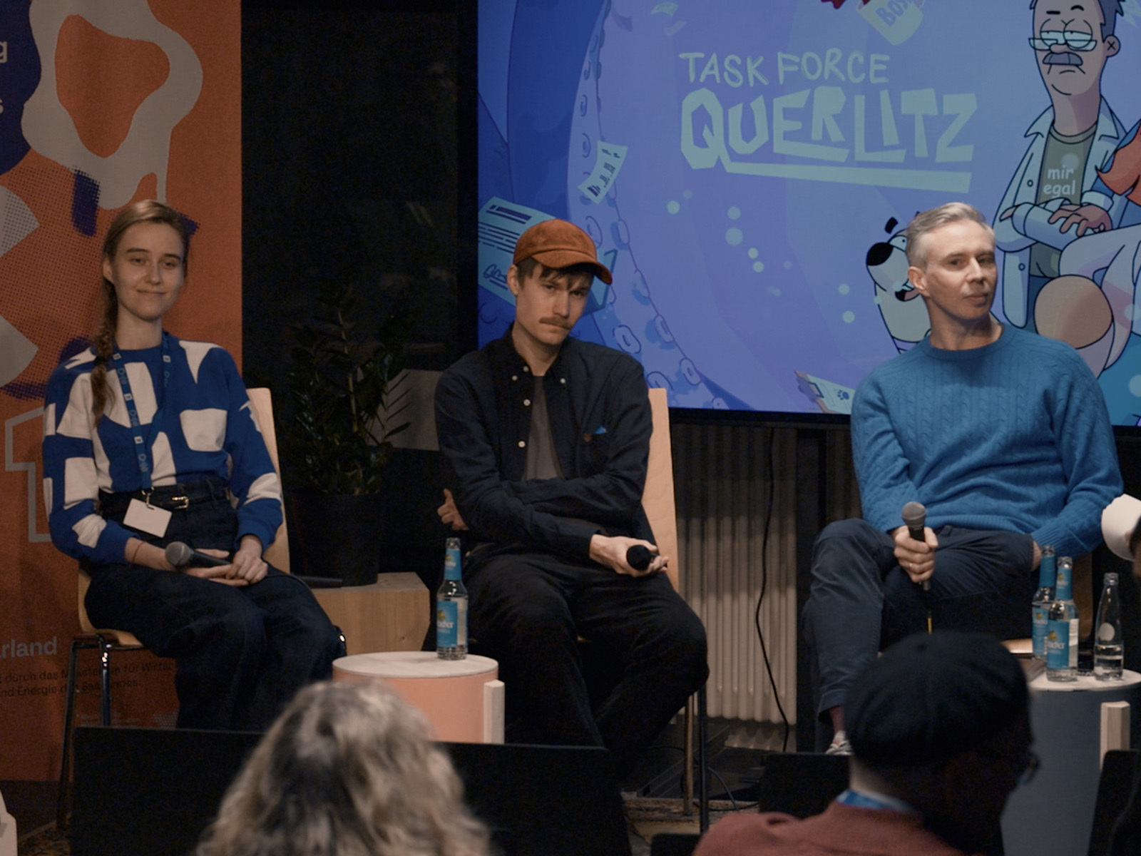 Podiumsdiskussion. Drei Personen sitzen auf einer Bühne mit Mikrofonen: links eine Person in blau-weißem gemustertem Pullover, in der Mitte eine Person mit brauner Cap und schwarzem Hemd, rechts eine Person in hellblauem Pullover. Im Hintergrund eine Leinwand mit dem Logo 'Task Force Querlitz' und Comic-Illustrationen in Blau- und Grüntönen. Im Vordergrund sind unscharf Köpfe von Zuschauenden zu sehen.