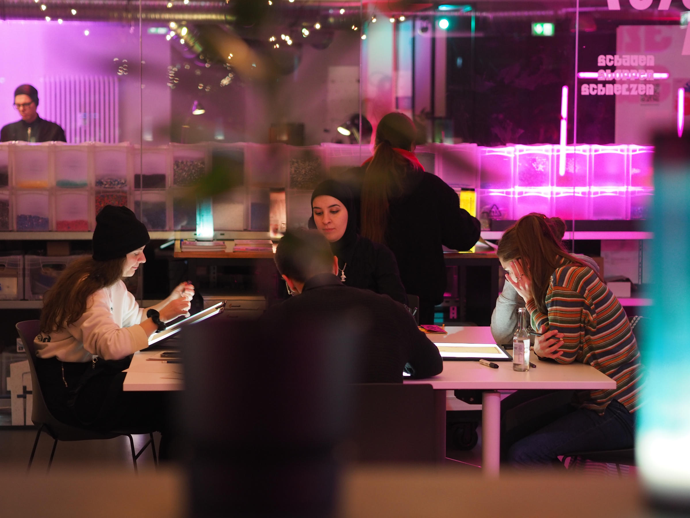 Networking-Situation bei einer Veranstaltung in einem modern beleuchteten Raum. Mehrere Personen sitzen an weißen Tischen in intensiv pinkem und türkisem Licht und führen Gespräche. Im Vordergrund verschwommene Silhouetten, im Hintergrund ein DJ-Pult, Bücherregale und eine Bar mit LED-Beleuchtung. Die Atmosphäre wirkt lebendig und kreativ.