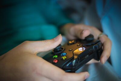 Nahaufnahme von Händen, die einen schwarzen Gaming-Controller halten. Deutlich sichtbar sind die beleuchteten Aktionstasten in Grün, Rot, Blau und Gelb sowie weitere Bedienelemente. Der Hintergrund ist in verschwommenen Blau- und Türkistönen gehalten, wodurch der Fokus auf dem Controller und der Gaming-Aktion liegt.