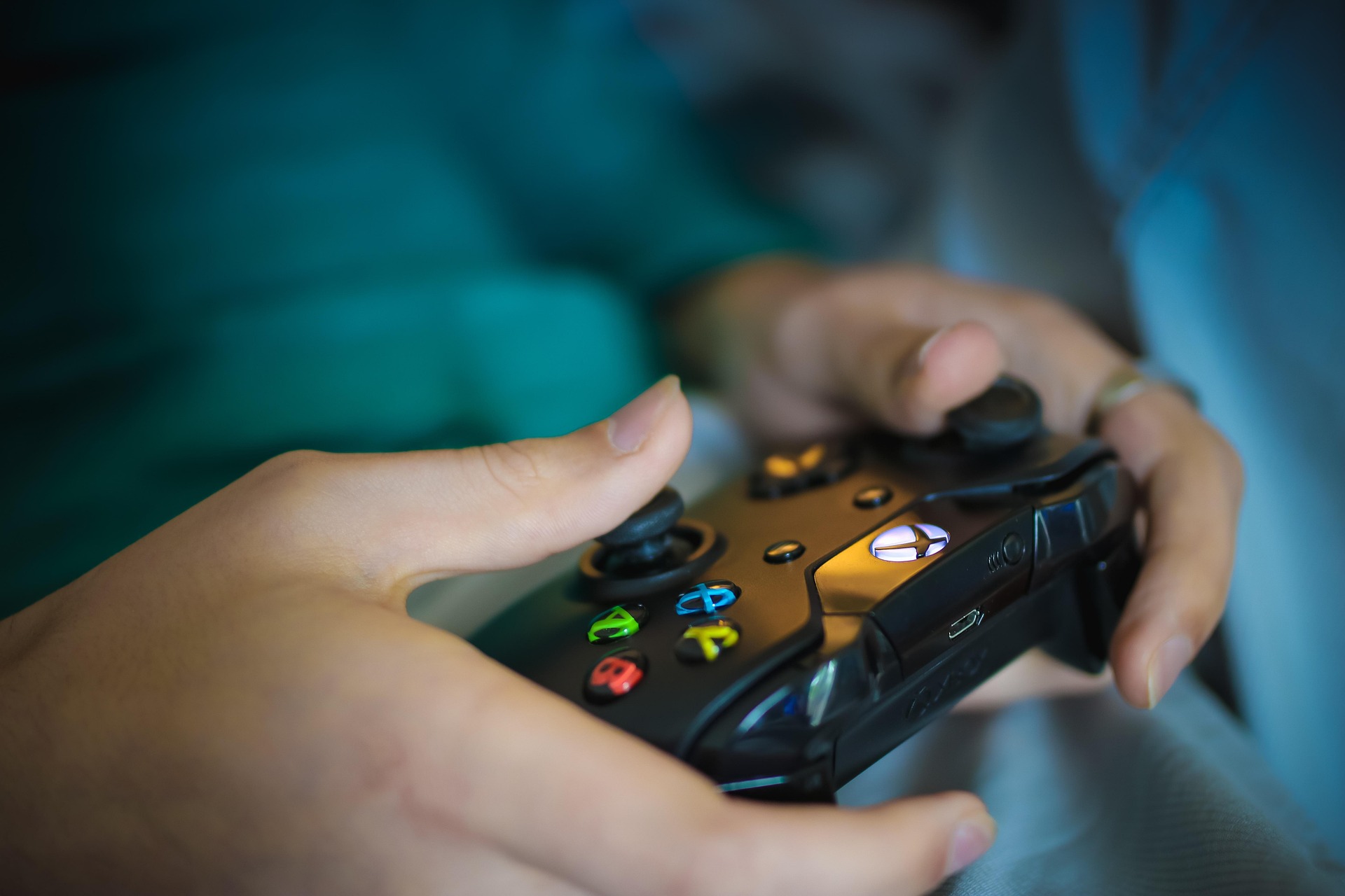 Nahaufnahme von Händen, die einen schwarzen Gaming-Controller halten. Deutlich sichtbar sind die beleuchteten Aktionstasten in Grün, Rot, Blau und Gelb sowie weitere Bedienelemente. Der Hintergrund ist in verschwommenen Blau- und Türkistönen gehalten, wodurch der Fokus auf dem Controller und der Gaming-Aktion liegt.