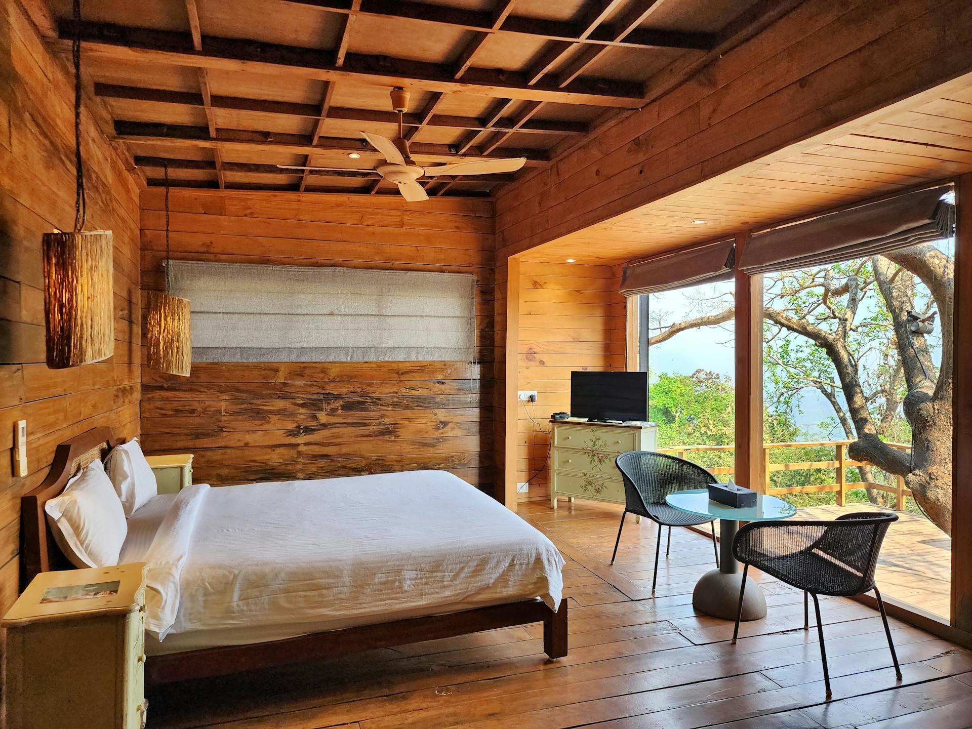 Hier ist ein Alt-Text für das Bild: "Luxuriöses Hotelzimmer in einer Holzblockhütte mit komplett holzverkleideten Wänden, Deckenbalken und Holzboden. Ein Doppelbett mit weißer Bettwäsche steht links, rustikale Holzlampen hängen von der Decke. Rechts eine kleine Sitzecke mit schwarzen modernen Stühlen und rundem Tisch vor einer großen Glasfront, die den Blick auf Bäume und eine grüne Landschaft freigibt. Eine Kommode mit Fernseher und ein Deckenventilator vervollständigen die warme, naturnahe Atmosphäre.