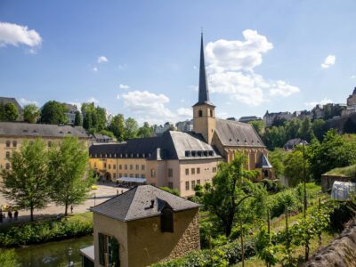 Malerische Stadtansicht von Luxemburg-Stadt an einem sonnigen Tag. Im Zentrum eine Kirche mit schlankem, hohem Kirchturm und dunkler Spitze, umgeben von historischen Gebäuden in sandfarbenen und ockerfarbenen Tönen. Grüne Bäume und Vegetation rahmen die Szenerie ein, im Hintergrund weitere Gebäude auf einem Hügel. Der blaue Himmel zeigt weiße Wolkenformationen.