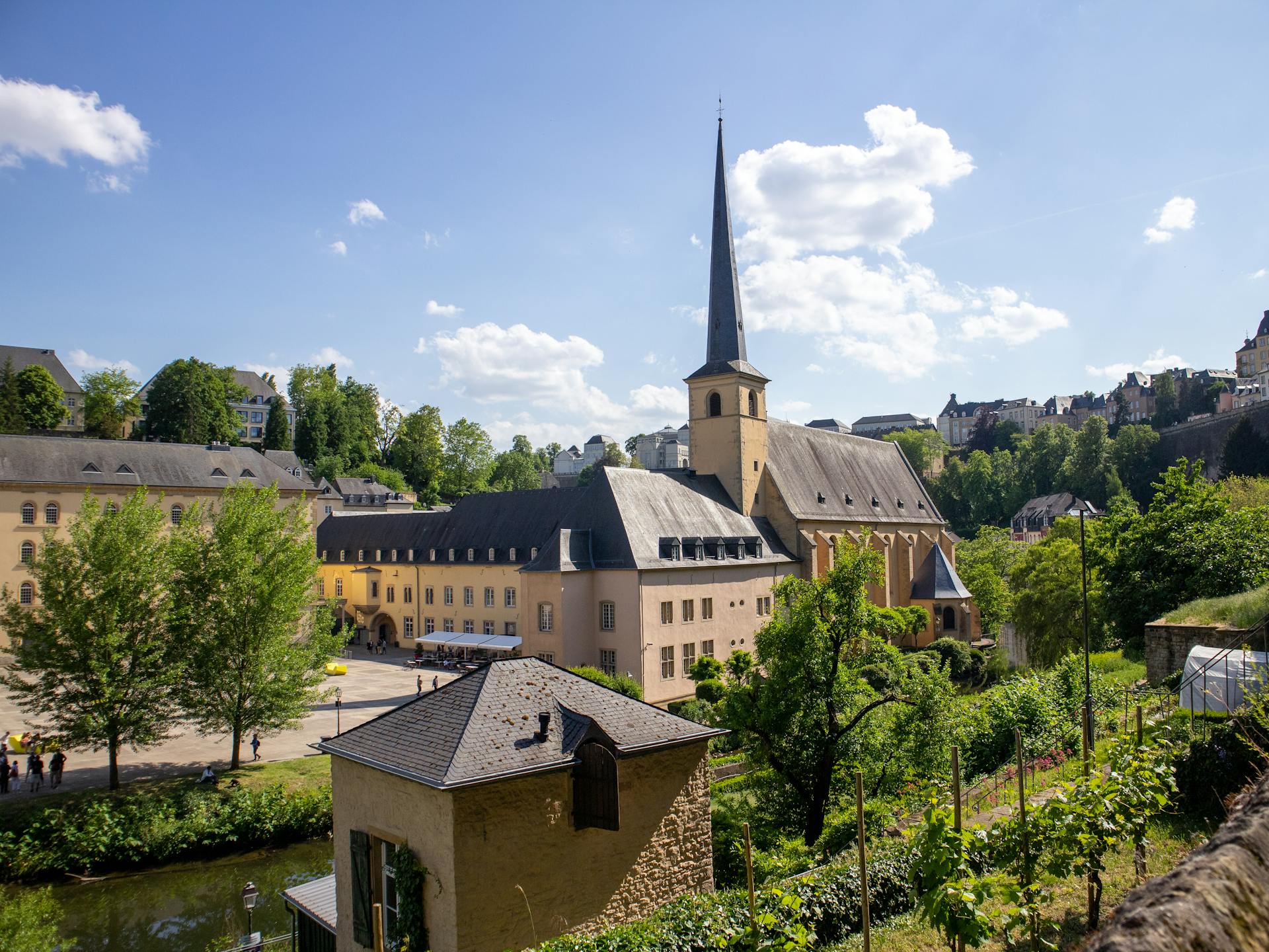 Malerische Stadtansicht von Luxemburg-Stadt an einem sonnigen Tag. Im Zentrum eine Kirche mit schlankem, hohem Kirchturm und dunkler Spitze, umgeben von historischen Gebäuden in sandfarbenen und ockerfarbenen Tönen. Grüne Bäume und Vegetation rahmen die Szenerie ein, im Hintergrund weitere Gebäude auf einem Hügel. Der blaue Himmel zeigt weiße Wolkenformationen.