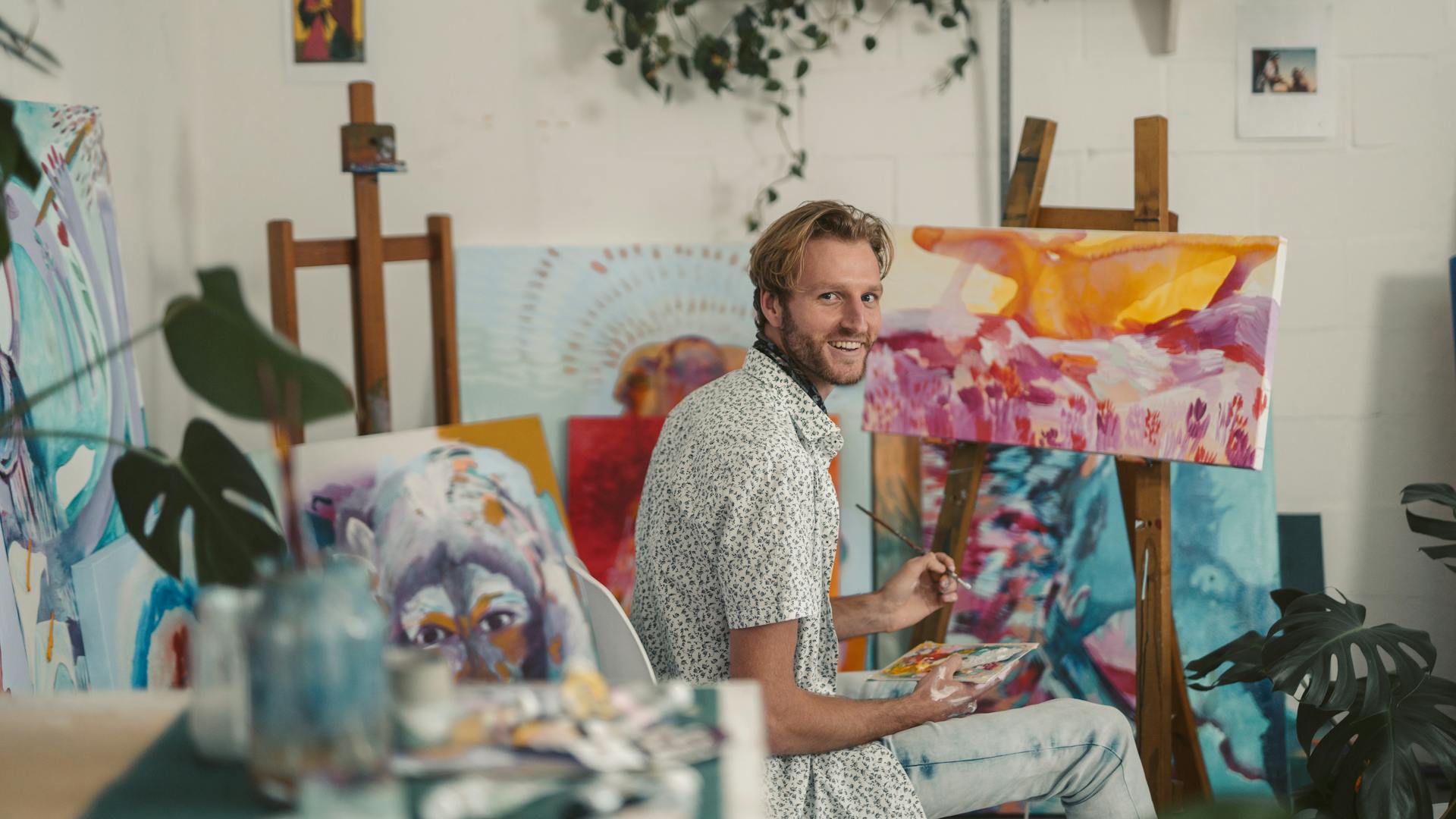 Ein Mann sitzt in einem Atelier vor einer Staffelei, hält Pinsel und Farbpalette in der Hand und blickt lächelnd zur Kamera; um ihn herum stehen mehrere farbintensive Gemälde und Pflanzen.