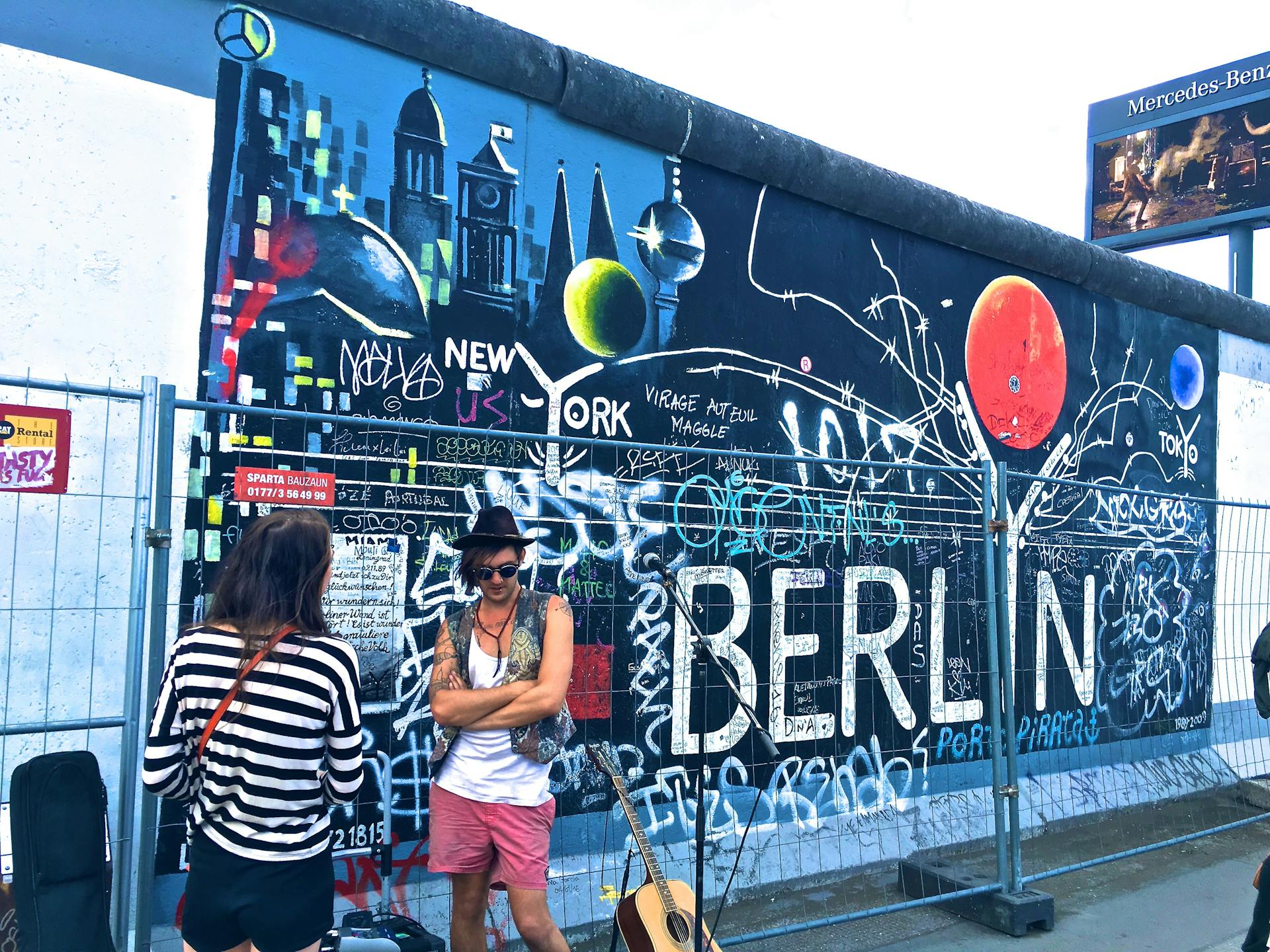 Zwei Touristen betrachten ein farbenfrohes Graffiti-Kunstwerk an einem erhaltenen Segment der Berliner Mauer. Das Mural zeigt Stadtsilhouetten, Weltkugeln und den großen Schriftzug 'BERLIN' sowie Referenzen zu Städten wie New York und Tokyo. Die Wand ist mit zahlreichen Graffiti-Tags und Kritzeleien übersät. Links eine Person in schwarz-weiß gestreiftem Shirt, rechts ein Mann mit Gitarre, Sonnenbrille und schwarzem Hut. Im Hintergrund weitere Mauerreste und ein Mercedes-Benz-Werbeschild.