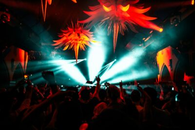 Innenansicht eines gut besuchten Nachtclubs oder Musikfestivals bei Nacht. Von der Decke hängen große, rot leuchtende, stachelige Dekorationen, die an abstrakte Sonnen oder Kreaturen erinnern. Helle, türkisfarbene Lichtstrahlen schneiden durch den dunklen Raum und beleuchten den Dunst in der Luft. Im Vordergrund ist die Silhouette einer tanzenden Menschenmenge zu sehen, viele mit erhobenen Armen, während einige den Moment mit ihren Smartphones filmen. Die Atmosphäre ist energiegeladen und kontrastreich.