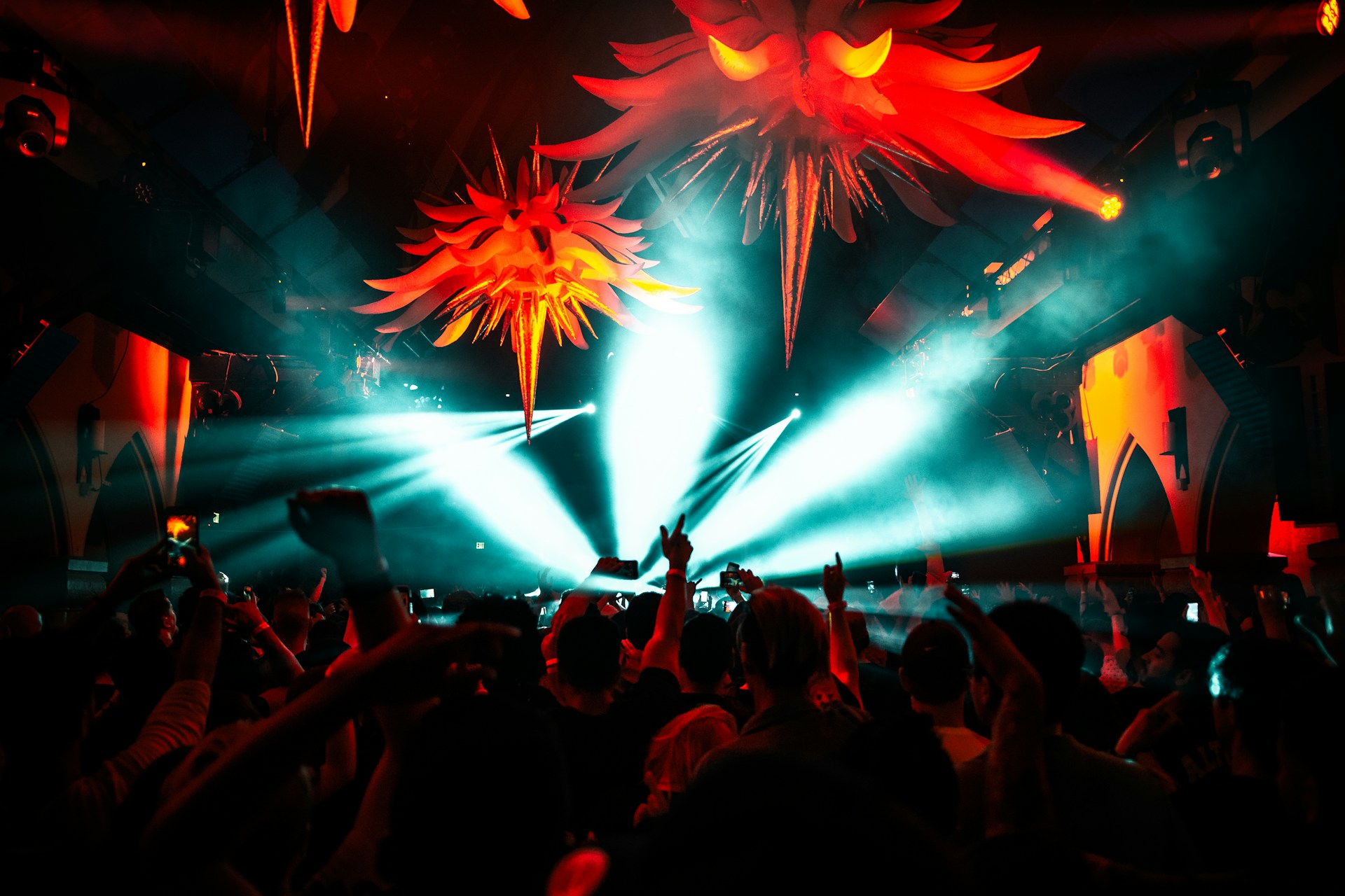Innenansicht eines gut besuchten Nachtclubs oder Musikfestivals bei Nacht. Von der Decke hängen große, rot leuchtende, stachelige Dekorationen, die an abstrakte Sonnen oder Kreaturen erinnern. Helle, türkisfarbene Lichtstrahlen schneiden durch den dunklen Raum und beleuchten den Dunst in der Luft. Im Vordergrund ist die Silhouette einer tanzenden Menschenmenge zu sehen, viele mit erhobenen Armen, während einige den Moment mit ihren Smartphones filmen. Die Atmosphäre ist energiegeladen und kontrastreich.
