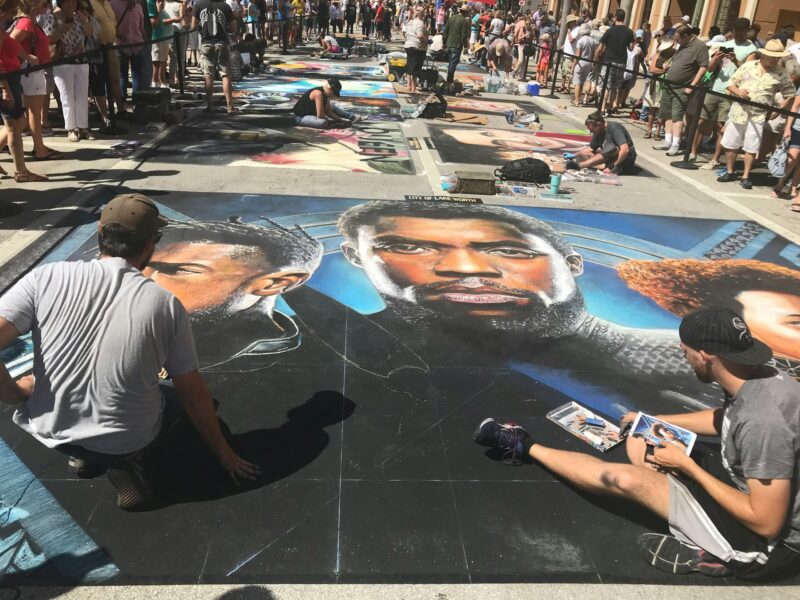 Eine Weitwinkelaufnahme eines Straßenkunst-Festivals auf hellem Asphalt. Im Vordergrund knien zwei Künstler und arbeiten an einem großformatigen, realistischen Kreideporträt von Charakteren aus dem Film Black Panther, darunter T'Challa im Zentrum. Die Zeichnung besticht durch lebendige Farben und starke Lichtkontraste. Im Hintergrund erstreckt sich die Straße mit weiteren Kunstwerken, an denen Künstler sitzen und arbeiten, während zahlreiche Zuschauer hinter Absperrbändern zusehen. Die Szene ist hell von der Sonne erleuchtet.