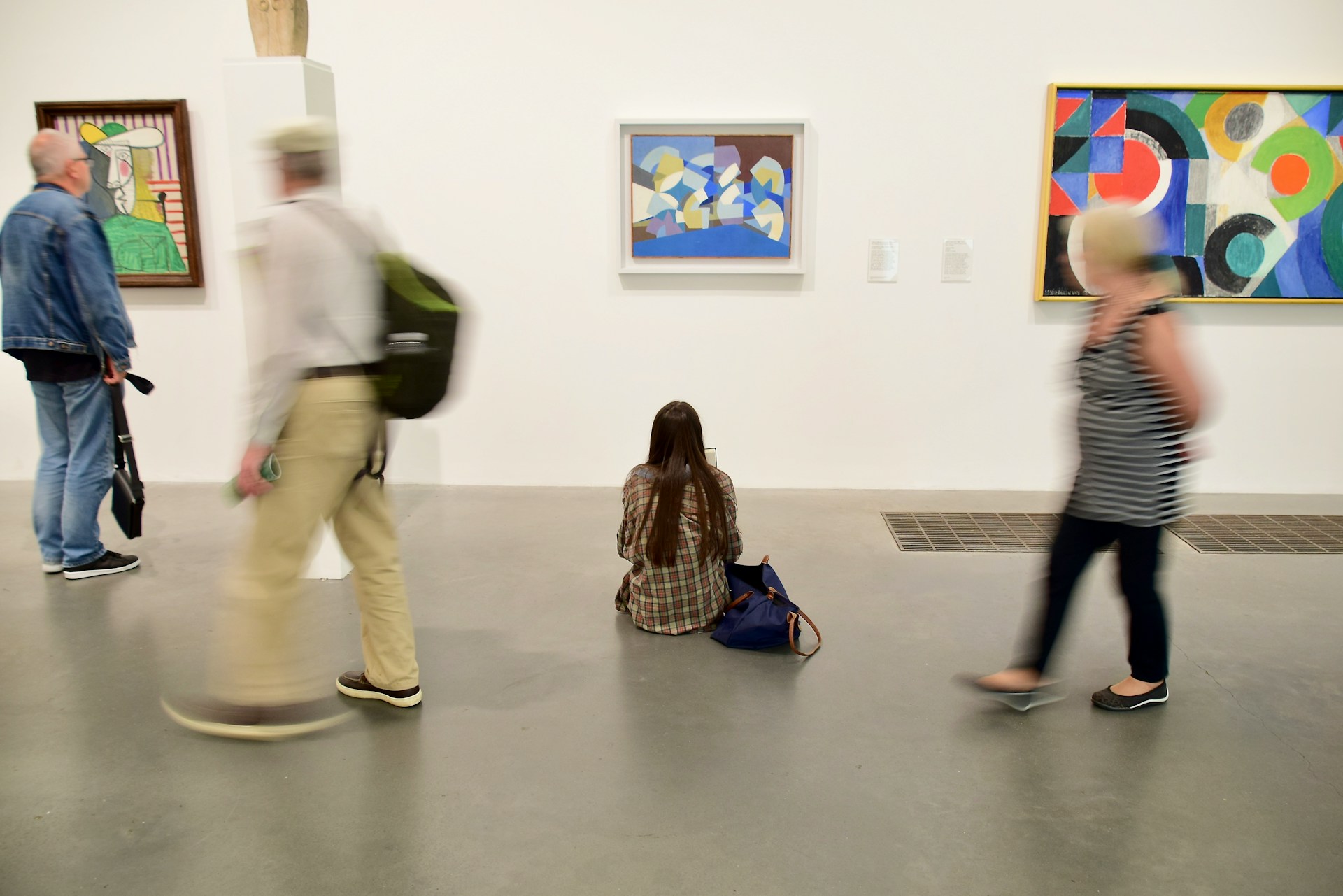 Museumsszene in einer Galerie für moderne Kunst. An weißen Wänden hängen drei bunte abstrakte Gemälde mit geometrischen Formen. Im Vordergrund sitzt eine Frau mit langen braunen Haaren in kariertem Hemd auf dem grauen Betonboden und betrachtet versunken die Kunstwerke. Um sie herum bewegen sich verschwommen weitere Besucherinnen und Besucher durch den Raum. Die Szene vermittelt einen Kontrast zwischen kontemplativer Ruhe und geschäftiger Bewegung.
