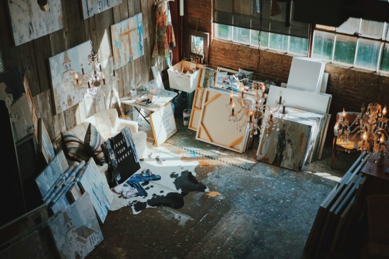 Künstleratelier in einem rustikalen Loft-Raum mit Backsteinwänden und großen Fenstern. Mehrere Staffeleien mit Leinwänden, teilweise fertige und unfertige Gemälde lehnen an den Wänden. Der Boden ist mit Farbspritzern bedeckt, überall verstreut Malutensilien, Pinsel und Farbtuben. Rechts hängen kristallene Kronleuchter, Sonnenlicht fällt durch die industriellen Fenster und schafft eine inspirierende, kreative Arbeitsatmosphäre.