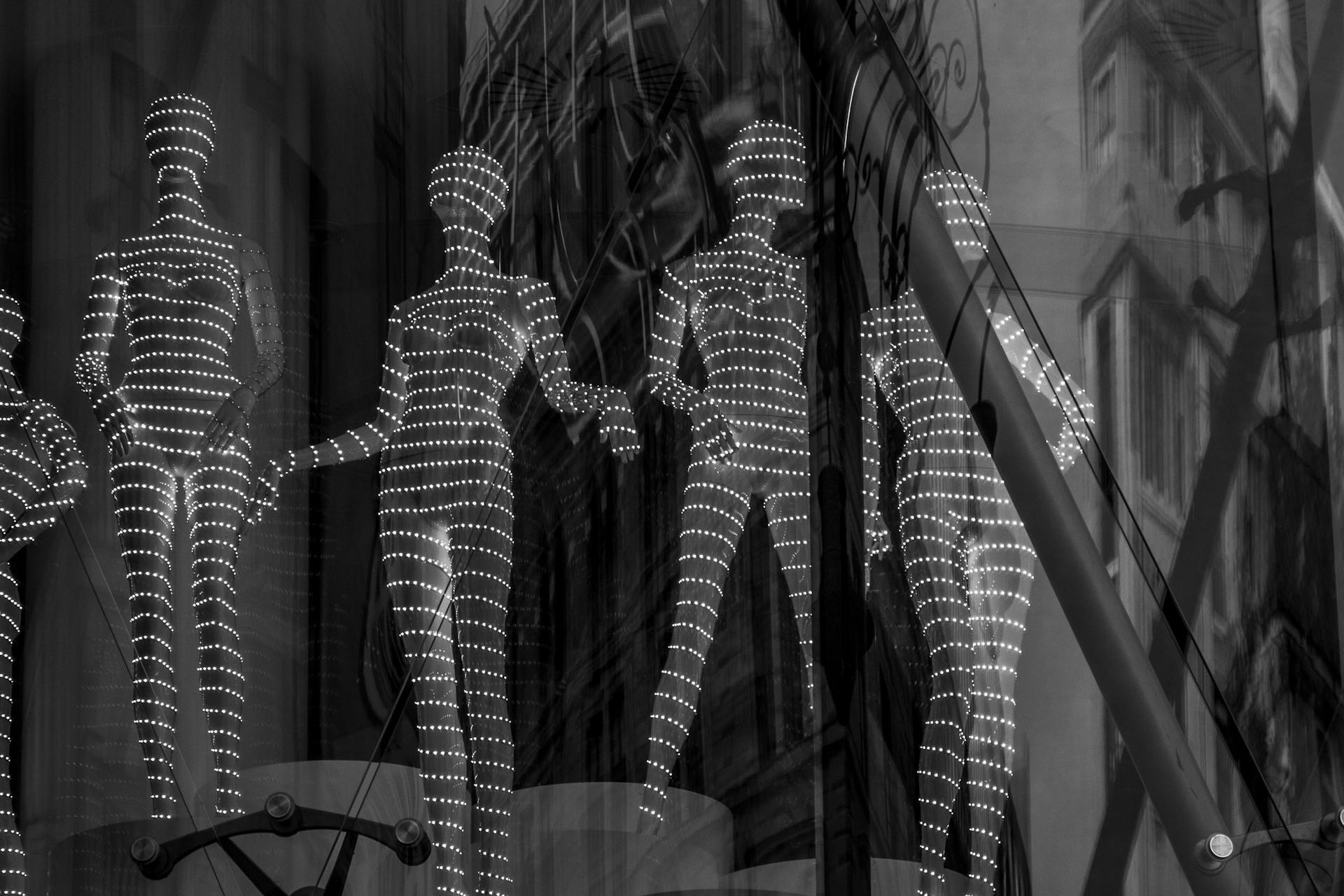 Schwarzweiß-Foto von Schaufensterpuppen in glitzernden Abendkleidern hinter einer Glasscheibe. Die Kleider sind mit feinen weißen Punktmustern in geschwungenen Linien verziert, die elegant über die dunklen Stoffe verlaufen. Durch Spiegelungen in der Scheibe überlagern sich die Reflexionen von Straßenszenen und Gebäuden mit den Figuren, was eine mehrschichtige, urbane Komposition erzeugt