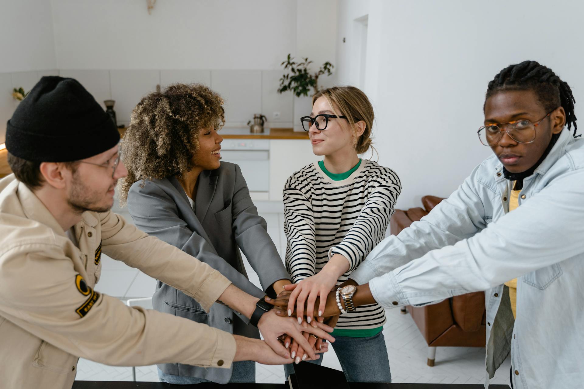 Vier junge Erwachsene stehen in einer Küche zusammen und legen ihre Hände übereinander, als Zeichen von Teamgeist und Zusammenarbeit.