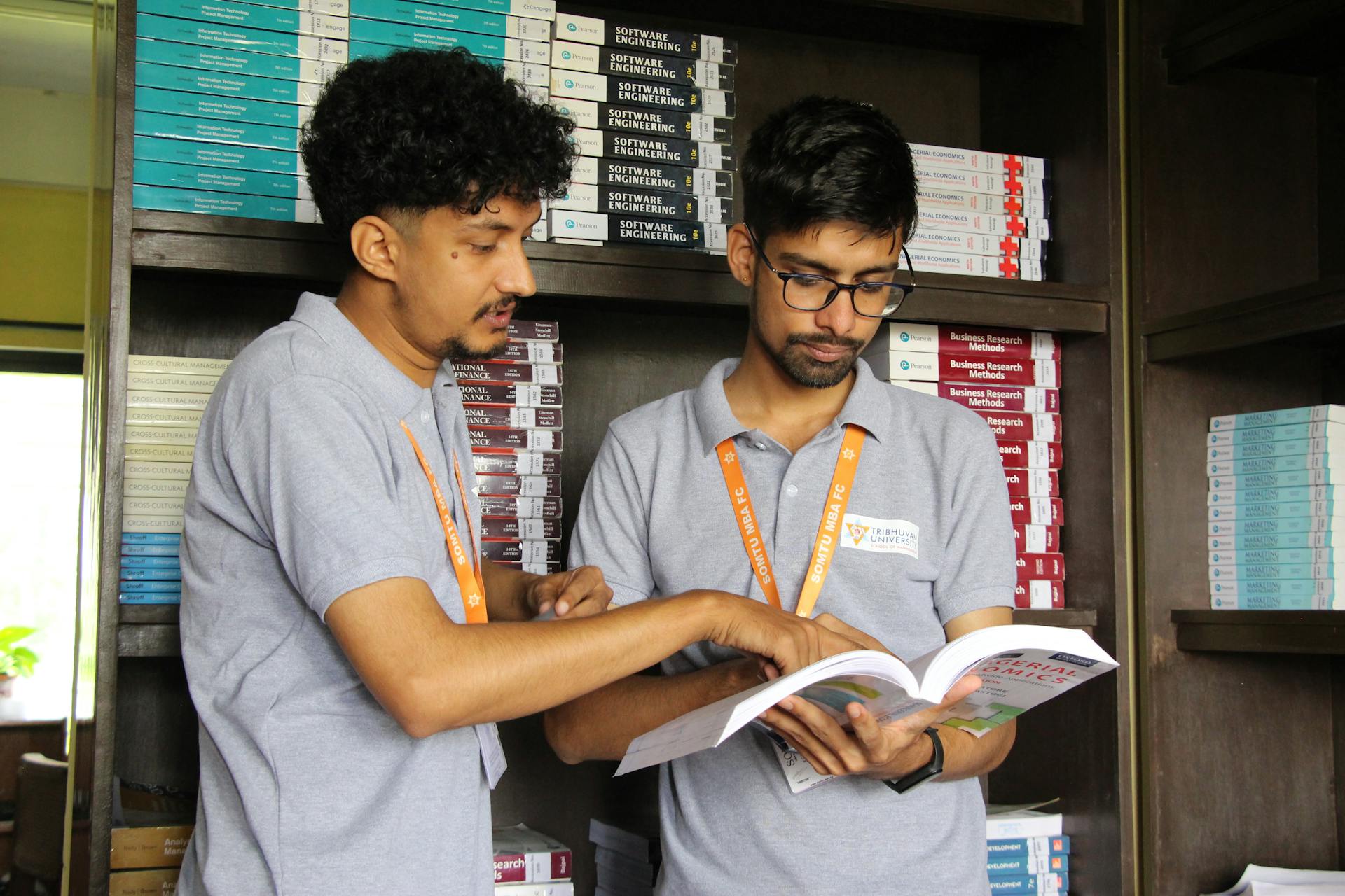 Zwei Studierende in grauen Poloshirts mit orangefarbenen Lanyards stehen in einer Bibliothek vor Bücherregalen und studieren gemeinsam ein aufgeschlagenes Buch. Der linke Student mit lockigen Haaren zeigt auf eine Stelle im Buch, während der rechte Student mit Brille konzentriert liest. Im Hintergrund gestapelte Lehrbücher zu Themen wie Software Engineering, Business Research Methods und Economics.