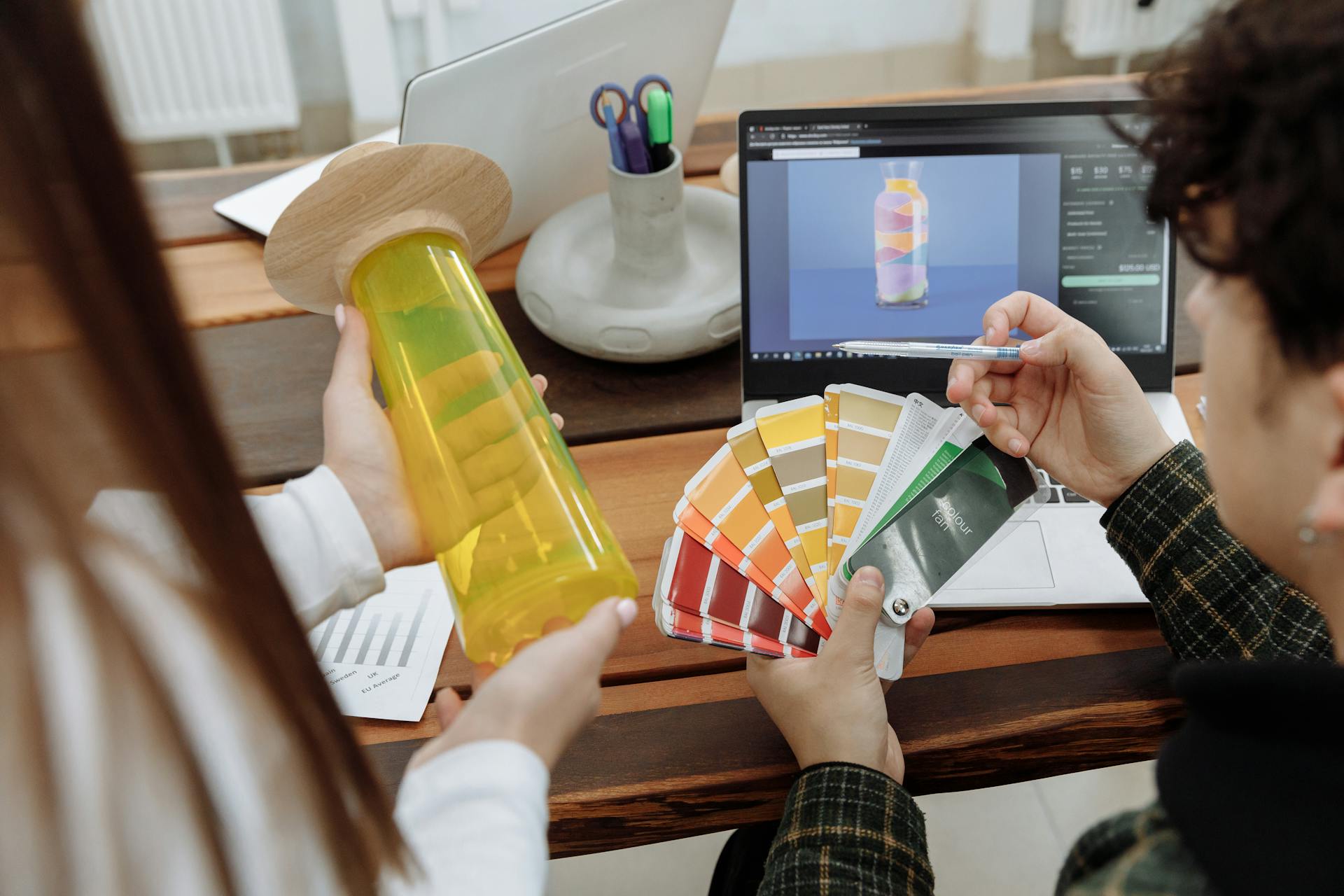 Produktdesign-Arbeitsplatz: Zwei Personen arbeiten gemeinsam an einem Holztisch. Eine Person hält einen gelb-grünen Prototyp einer Flasche oder eines Behälters, die andere Person hält einen Farbfächer mit verschiedenen Farbmustern. Auf dem Laptop-Bildschirm im Hintergrund ist ein 3D-Rendering desselben Flaschendesigns in ähnlichen Farben zu sehen. Der Arbeitsplatz ist mit Design-Materialien, einem weißen iMac und Büromaterialien ausgestattet. Die Szene zeigt den kreativen Prozess von der digitalen Gestaltung bis zum physischen Prototyp.