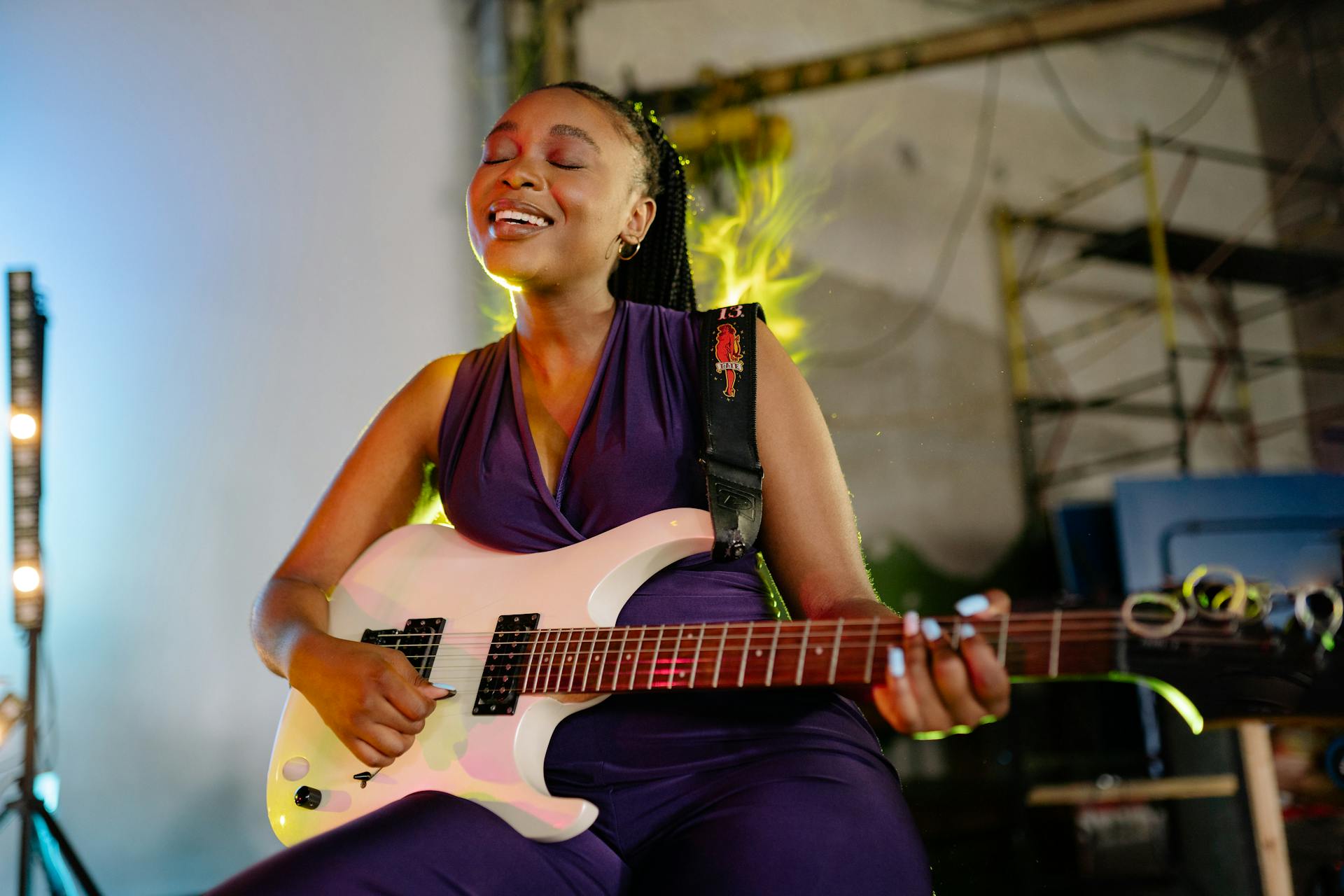 Eine junge Frau mit geschlossenen Augen und einem glücklichen Lächeln spielt eine weiße E-Gitarre. Sie trägt einen violetten Jumpsuit und steht in einem stimmungsvoll beleuchteten Raum, der an ein Studio oder eine Bühne erinnert. Warmes, gelbes Licht hebt ihre Silhouette hervor und erzeugt eine dynamische, musikalische Atmosphäre.