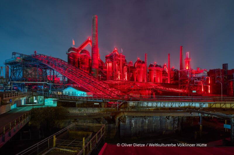 Hier ist ein Alt-Text für das Bild: "Nachtaufnahme der Völklinger Hütte, UNESCO-Weltkulturerbe, in spektakulärer Beleuchtung. Die monumentale Hochofengruppe und die gesamte Industrieanlage sind in intensives rotes Licht getaucht, das die massiven Öfen, Schornsteine, Förderbänder und die komplexe Stahlkonstruktion dramatisch hervorhebt. Links türkisblaue Beleuchtungsakzente auf Gebäudeteilen. Der dunkelblaue Abendhimmel bildet einen eindrucksvollen Kontrast zur rot leuchtenden Industriearchitektur. Im Vordergrund Wasseranlagen und Rohrleitungen des ehemaligen Stahlwerks. Die künstlerische Lichtinszenierung verwandelt das Industriedenkmal in ein beeindruckendes nächtliches Spektakel.