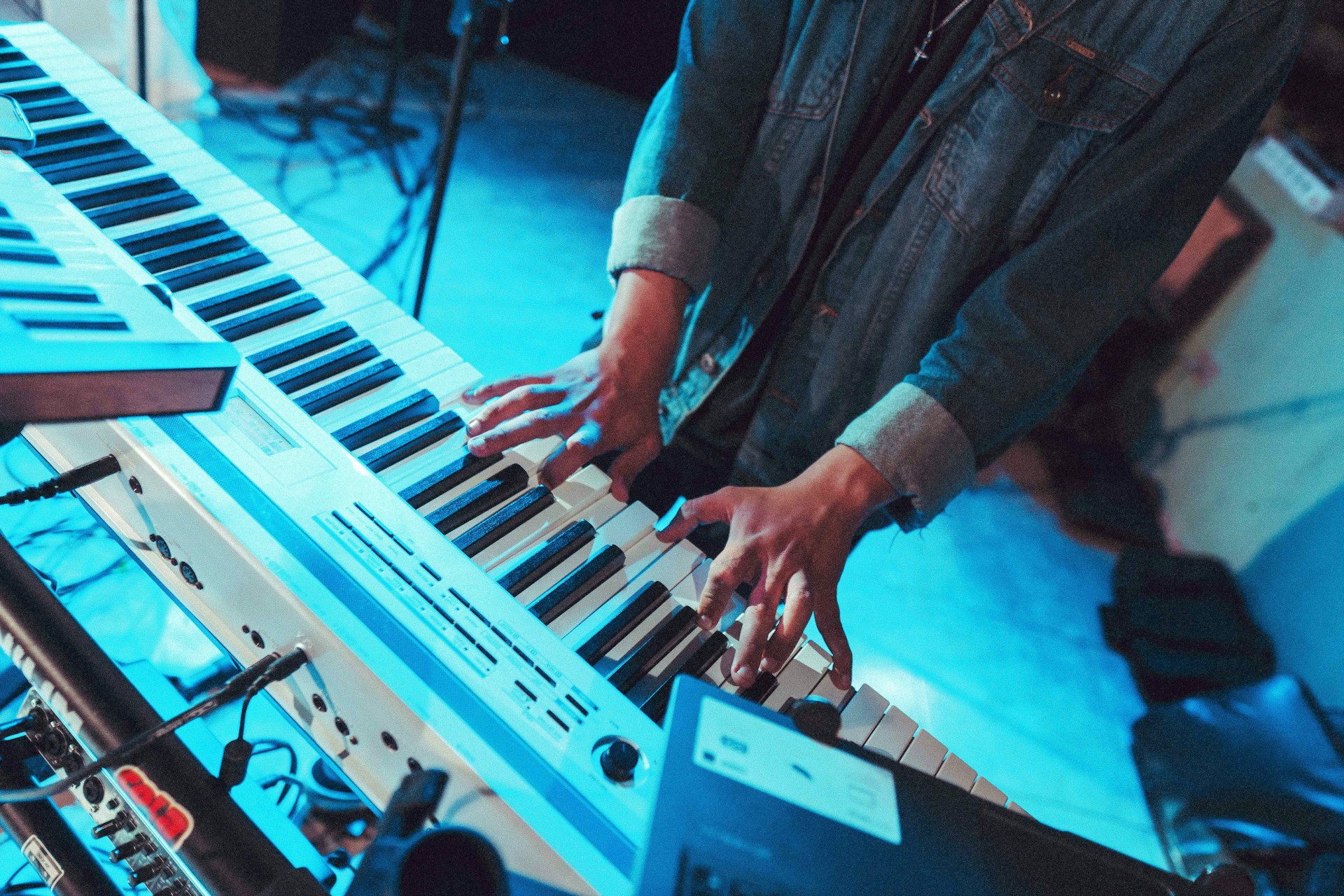 Nahaufnahme eines Musikers an einem elektronischen Keyboard in atmosphärischem blauen Bühnenlicht. Die Hände spielen auf den schwarz-weißen Tasten des Synthesizers. Die Person trägt eine dunkle Jeansjacke. Im Vordergrund sind weitere Musiker unscharf erkennbar, im Hintergrund Kabel und weiteres Musikequipment. Die Szene vermittelt die Atmosphäre einer Live-Performance oder Studioaufnahme.