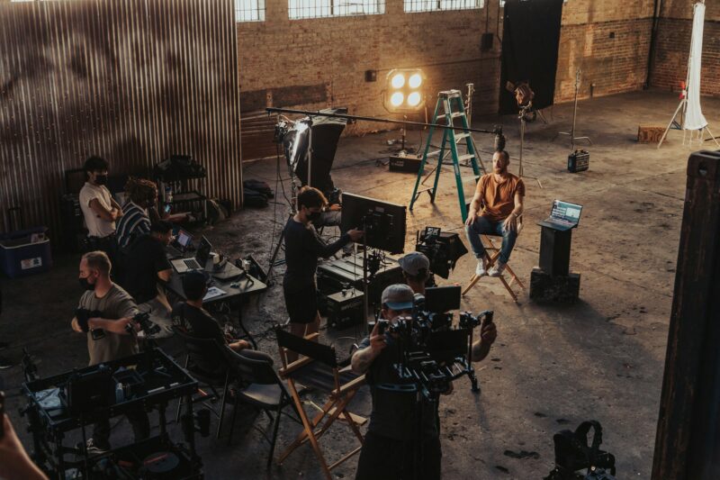 Professionelles Filmset in einer großen verlassenen Industriehalle mit Backsteinwänden und hohen vergitterten Fenstern. Eine diverse Filmcrew von etwa zehn Personen arbeitet an einer Szene: Ein Schauspieler in orangefarbenem Hemd sitzt auf einem Regiestuhl vor der Kamera, während Kameramänner, Ton- und Lichttechniker um ihn herum positioniert sind. Mehrere Laptops, ein türkises Gerüst mit Lampen und professionelle Filmausrüstung sind sichtbar. Die natürliche Beleuchtung von den großen Fenstern erzeugt warme Lichter in der ansonsten dunklen, räumlichen Halle. Die Szene zeigt einen konzentrierten Filmproduktionsprozess.