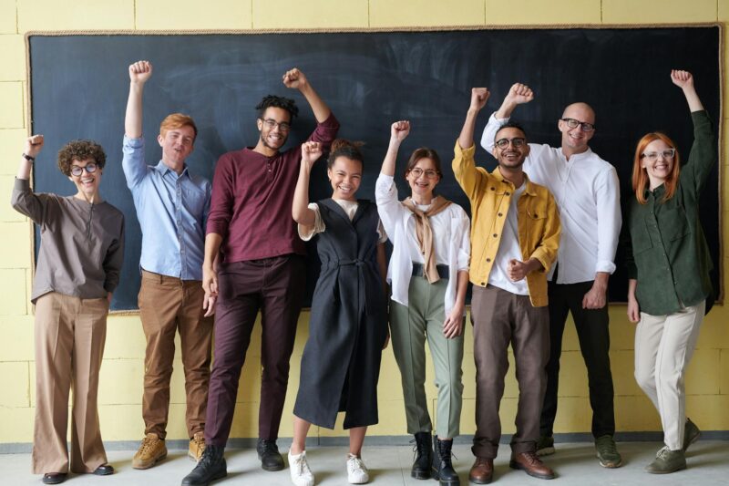 Acht fröhliche Teammitglieder stehen in einer Reihe vor einer großen schwarzen Tafel und einer gelben Wand. Alle recken begeistert die Fäuste in die Höhe – eine Erfolgsgeste. Das diverse Team trägt legere Business-Kleidung in verschiedenen gedeckten Farben: Grau, Blau, Bordeaux, Schwarz, Weiß, Gelb und Grün. Mehrere Personen tragen Brillen. Die Atmosphäre ist fröhlich und motiviert, die Körpersprache vermittelt gemeinsamen Erfolg und Teamgeist. Die Szene könnte ein erfolgreiches Projekt, einen Workshop-Abschluss oder einen Team-Meilenstein feiern.