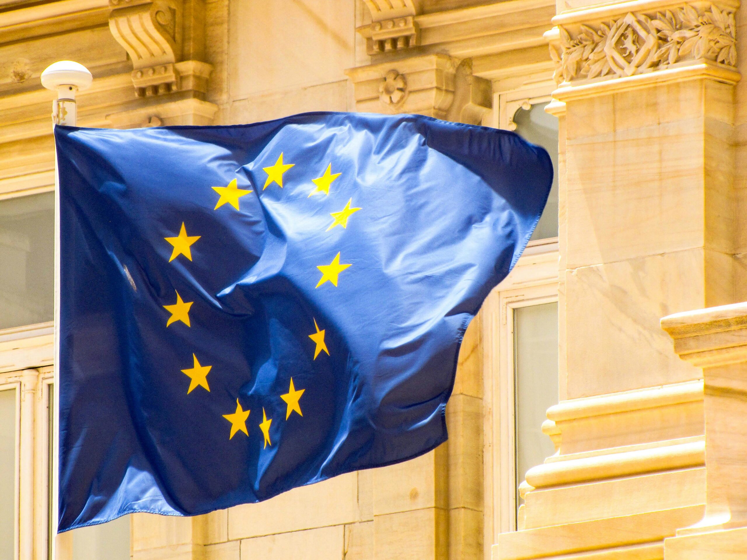 Die Flagge der Europäischen Union weht vor einem prächtigen historischen Gebäude mit beiger Sandsteinfassade und aufwendigen klassizistischen Verzierungen. Die blaue Flagge zeigt den charakteristischen Kreis aus zwölf goldenen Sternen. Im Hintergrund kunstvoll gearbeitete Säulen, Kapitelle und Ornamente, die von warmem Licht angestrahlt werden. Die Szene vermittelt europäische Institutionen und historische Architektur.