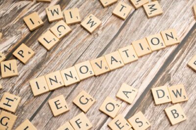 Scrabble-Buchstabensteine auf verwittertem Holzuntergrund. In der Bildmitte deutlich lesbar das Wort 'INNOVATION', horizontal gelegt aus hellen Holzbuchstaben mit schwarzen Buchstaben und kleinen Punktezahlen. Rundherum verstreut weitere Buchstabensteine, teilweise zu unlesbaren Fragmenten angeordnet. Die rustikale Holzmaserung bildet einen natürlichen, warmen Hintergrund. Das Bild symbolisiert Kreativität, Ideenfindung und spielerisches Denken.