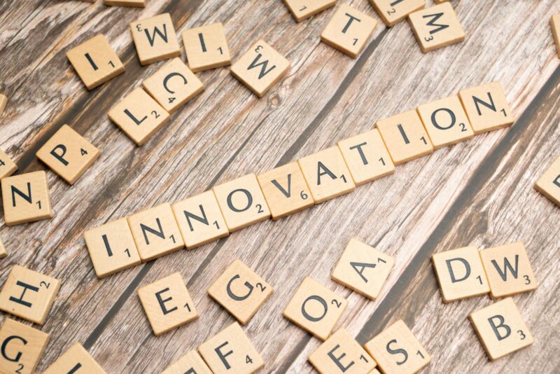 Scrabble-Buchstabensteine auf verwittertem Holzuntergrund. In der Bildmitte deutlich lesbar das Wort 'INNOVATION', horizontal gelegt aus hellen Holzbuchstaben mit schwarzen Buchstaben und kleinen Punktezahlen. Rundherum verstreut weitere Buchstabensteine, teilweise zu unlesbaren Fragmenten angeordnet. Die rustikale Holzmaserung bildet einen natürlichen, warmen Hintergrund. Das Bild symbolisiert Kreativität, Ideenfindung und spielerisches Denken.