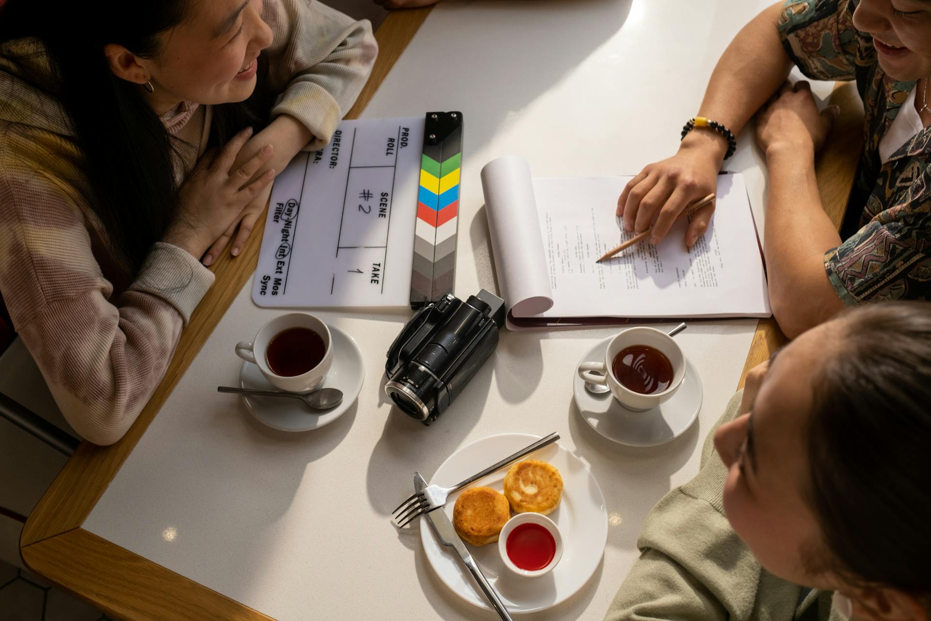 Vogelperspektive auf drei Personen bei einer Filmbesprechung an einem weißen Tisch. Eine bunte Filmklappe mit den Feldern 'DIRECTOR', 'SCENE', 'TAKE', 'ROLL' liegt neben einer professionellen Videokamera. Eine Person rechts zeigt mit einem Stift auf ein aufgeschlagenes Drehbuch oder Skript. Auf dem Tisch stehen zwei Tassen Tee und ein Teller mit zwei kleinen runden Gebäckstücken und roter Sauce. Die Atmosphäre vermittelt eine kreative Filmproduktions-Besprechung.
