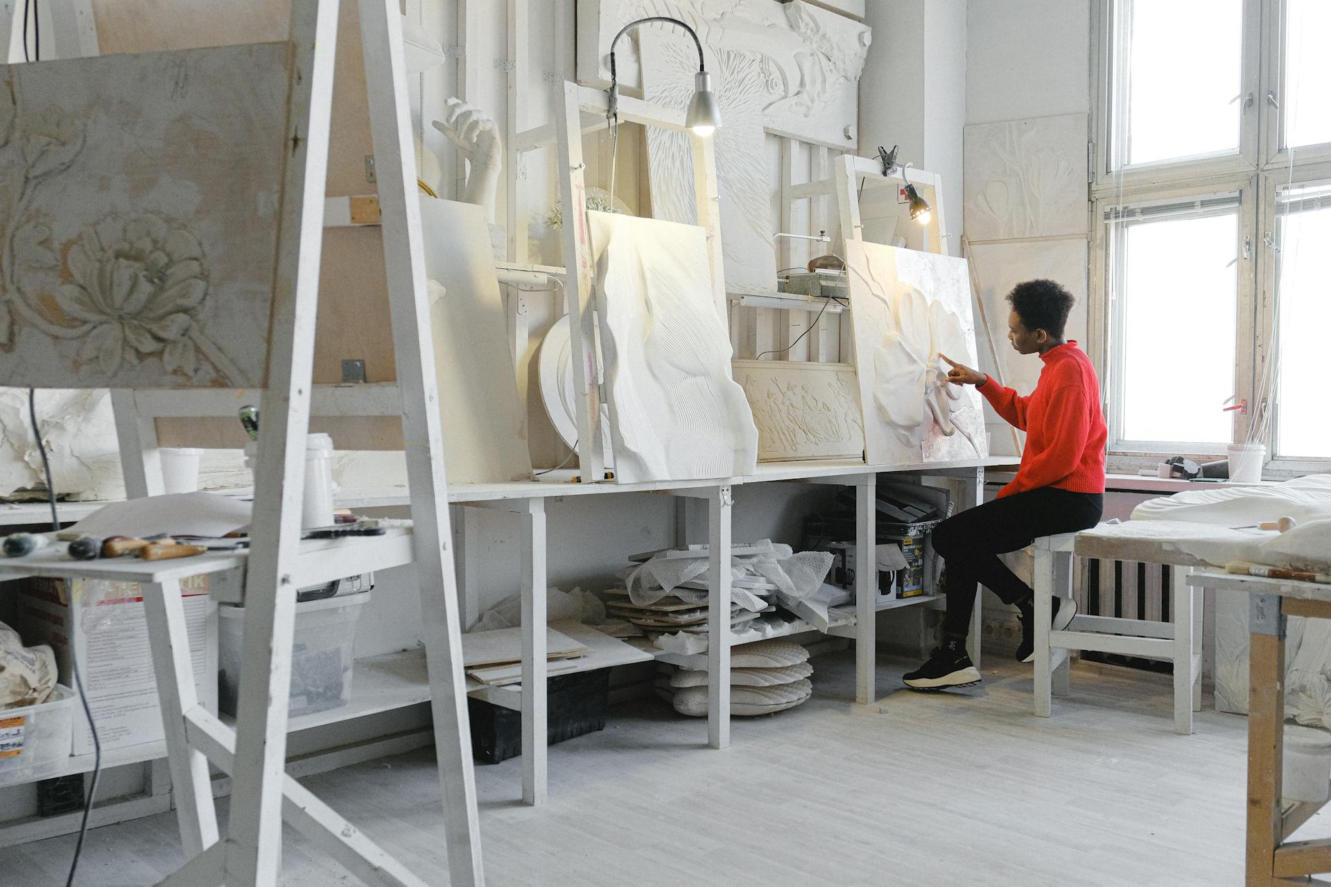 Künstlerin in einem hellen Bildhauer-Atelier bei der Arbeit an einer großformatigen abstrakten Leinwand. Sie trägt eine leuchtend orangefarbene Jacke und sitzt auf einer weißen Bank vor ihrem Werk, den Pinsel in der Hand. Das lichtdurchflutete Atelier mit hohen Fenstern und weißen Wänden ist angefüllt mit Gipsabgüssen, Reliefs und Skulpturen auf weißen Regalen und Arbeitsflächen. Links eine Leiter, im gesamten Raum kreatives Chaos aus Materialien und unfertigen Kunstwerken. Die Atmosphäre ist hell, inspirierend und produktiv.