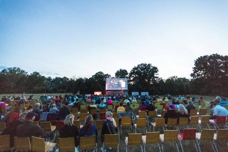 Open-Air-Kino in der Abenddämmerung auf einer Wiese. Zahlreiche Zuschauerinnen und Zuschauer sitzen in bunten Klappstühlen – überwiegend in Gelb, Rot und anderen Farbtönen – und schauen auf eine große Leinwand, auf der ein Film läuft. Im Hintergrund eine Baumreihe, die sich gegen den blauen Abendhimmel abzeichnet. Die Atmosphäre ist entspannt und sommerlich, die Beleuchtung wird bereits dämmrig. Ein typisches Sommerkino-Erlebnis unter freiem Himmel.