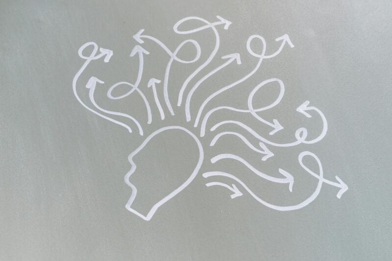 Grafische Illustration auf grauem Hintergrund: Ein vereinfachtes Profil eines menschlichen Kopfes in weißer Konturzeichnung, aus dem mehrere gebogene Pfeile in verschiedene Richtungen ausstrahlen. Die Pfeile sind mit Geschlechtssymbolen (♀ und ♂) verziert und symbolisieren vielfältige Gedanken, Ideen und unterschiedliche Perspektiven. Die Illustration repräsentiert Kreativität, Diversität, Gedankenaustausch und die Vielfalt menschlicher Gedanken und Überzeugungen.