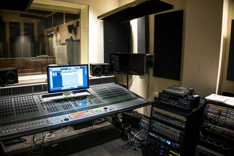 Professionelles Tonstudio mit Aufnahme- und Mischraum. Im Vordergrund ein großes digitales Mischpult mit zahlreichen Fadern, Knöpfen und bunten Tasten, dahinter ein Computer-Monitor mit Audio-Software. Links ein Fenster mit Blick in die schallisolierte Aufnahmekabine, rechts mehrere hochwertige Studio-Monitore (Lautsprecher) und gestapelte Rack-Geräte für Audio-Processing. Im Hintergrund große schwarze Akustik-Absorber an den Wänden. Die Beleuchtung ist warm und professionell, die Atmosphäre typisch für ein modernes Recording-Studio.