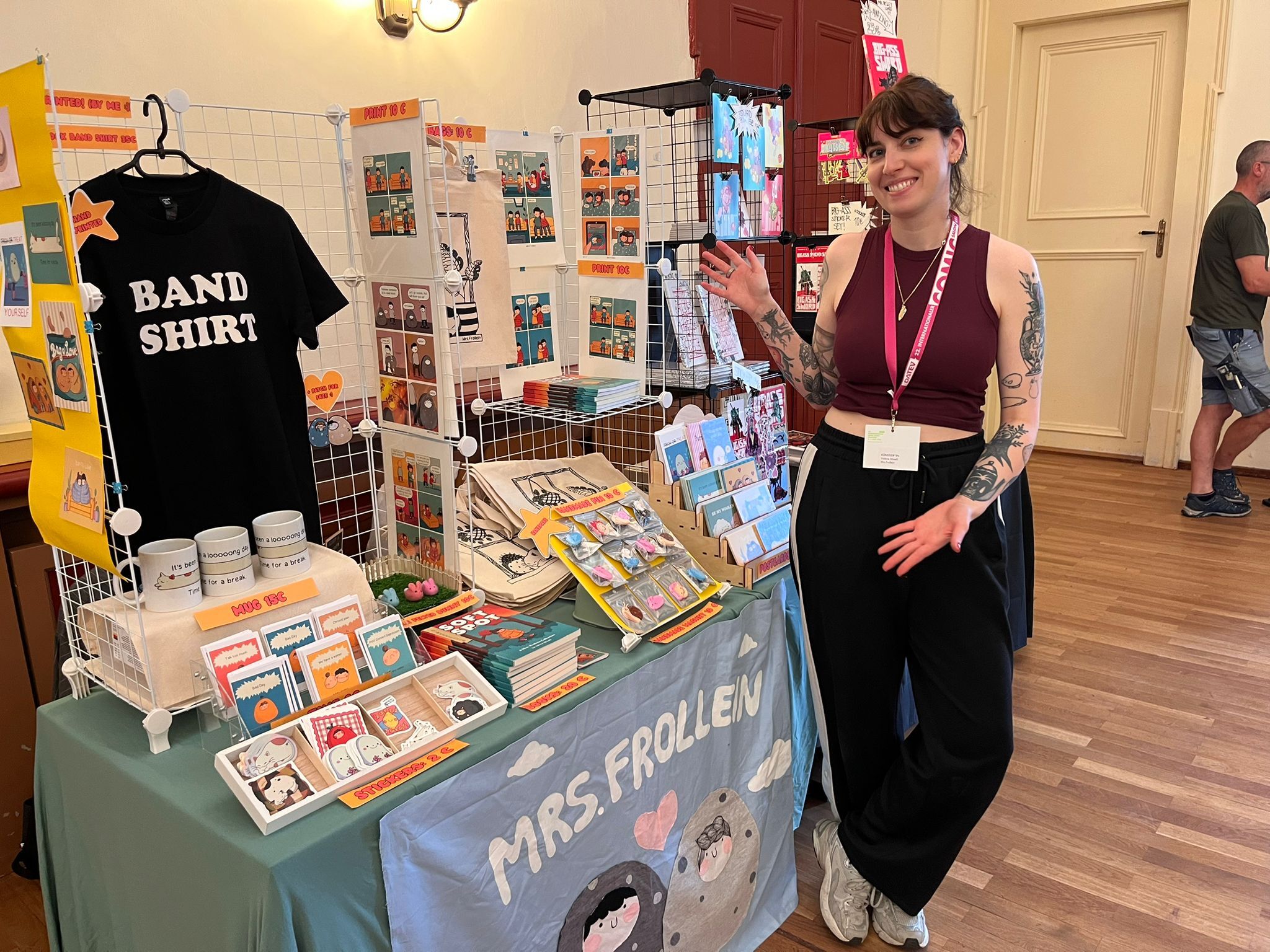 Comiczeichnerin Valérie Minelli mit Tattoos und dunklen Haaren steht lächelnd neben einem Verkaufsstand auf einer Comic- oder Kunstmesse. Sie trägt ein burgunderrotes Crop-Top, eine schwarze weite Hose und ein Lanyard mit Ausweis. Der Stand trägt den Namen „Mrs. Frollein" und zeigt eine Vielzahl selbstgemachter Produkte: Comics und Drucke, Sticker, Tassen mit der Aufschrift „Mug 15€", Tote Bags, Pins sowie ein schwarzes T-Shirt mit der Aufschrift „Band Shirt". Die Preisschilder zeigen Preise von 10 bis 15 Euro. Im Hintergrund sind weitere Messebesucher und ein Ausstellungsraum mit Parkettboden zu sehen.