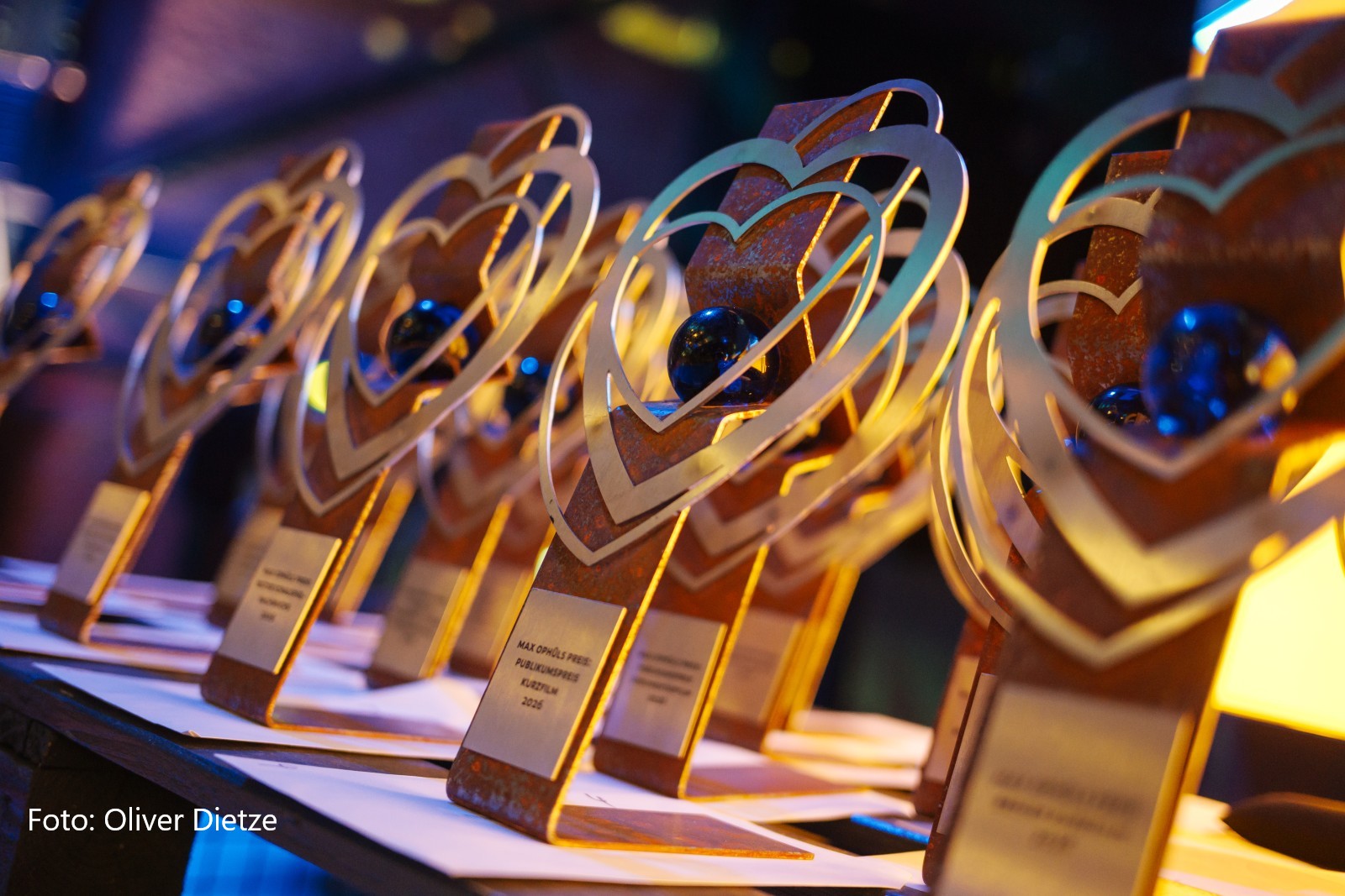 Nahaufnahme mehrerer Filmpreis-Trophäen des Max-Ophüls-Preises, aufgereiht auf einem Tisch. Die Trophäen bestehen aus stilisierten, herzförmigen Metallformen mit rostigem Sockel, in deren Mitte jeweils eine blaue Glaskugel eingebettet ist. Auf einer der gut sichtbaren Plaketten im Vordergrund steht „MAX OPHÜLS PREIS PUBLIKUMSPREIS KURZFILM 2026". Die Szene ist stimmungsvoll in warmes, golden-blaues Licht getaucht. Unten links im Bild der Bildnachweis „Foto: Oliver Dietze".