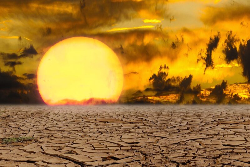 Alt-Text: Dramatische Bildkomposition zum Thema Klimawandel und Dürre: Im Vordergrund erstreckt sich ausgetrockneter, rissiger Lehmboden. Im Hintergrund geht eine überdimensional große, glutrote Sonne unter einem bedrohlich wirkenden, orangegelben Himmel mit dunklen Wolken und Dunst unter. Rechts sind schemenhaft Bäume zu erkennen. Das Bild vermittelt eine apokalyptische Stimmung und symbolisiert extreme Hitze und Wasserknappheit.