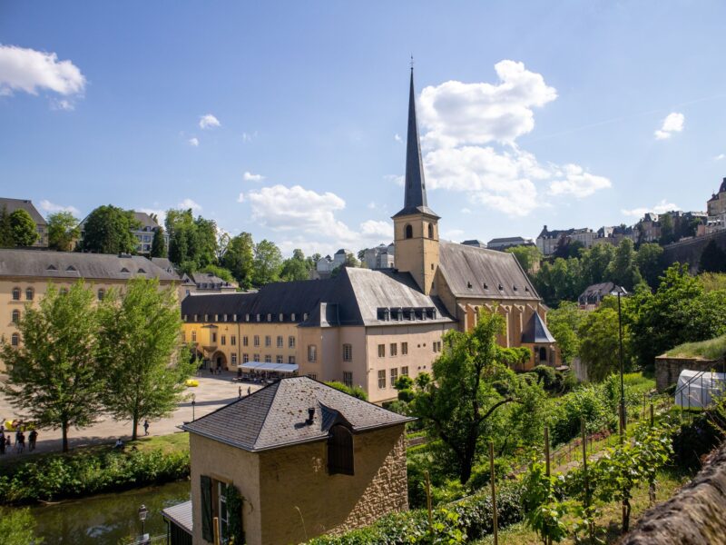 Alt-Text: Blick auf das historische Viertel Grund in Luxemburg-Stadt mit der Abtei Neumünster, deren Kirchturm mit schlankem, spitzem Turmhelm sich vor blauem Himmel mit weißen Wolken erhebt. Davor liegt ein kleines steinernes Gebäude mit Schieferdach. Die Umgebung ist von viel Grün geprägt, mit Bäumen, einem Weinberg sowie weiteren historischen Gebäuden. Im Hintergrund sind oberhalb der Schlucht weitere Häuser der Oberstadt zu sehen.