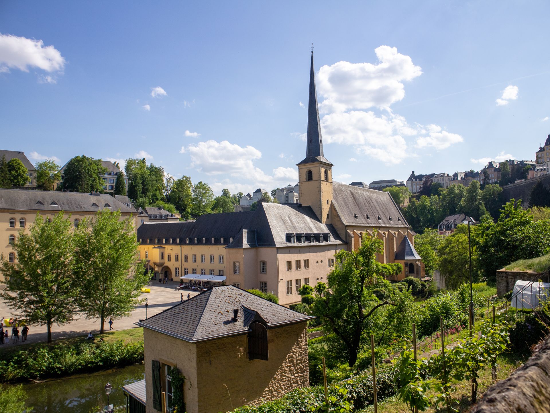 Alt-Text: Blick auf das historische Viertel Grund in Luxemburg-Stadt mit der Abtei Neumünster, deren Kirchturm mit schlankem, spitzem Turmhelm sich vor blauem Himmel mit weißen Wolken erhebt. Davor liegt ein kleines steinernes Gebäude mit Schieferdach. Die Umgebung ist von viel Grün geprägt, mit Bäumen, einem Weinberg sowie weiteren historischen Gebäuden. Im Hintergrund sind oberhalb der Schlucht weitere Häuser der Oberstadt zu sehen.