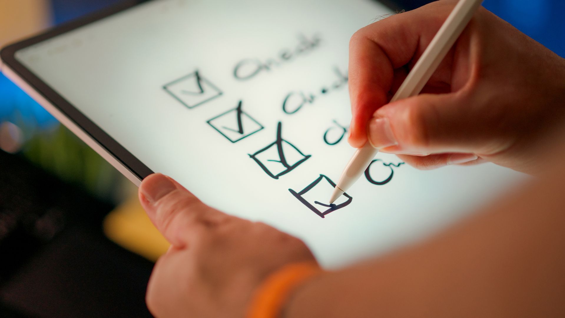 Nahaufnahme von zwei Händen, die ein Tablet halten. Eine Hand führt einen Eingabestift und hakt dabei handschriftlich gezeichnete Checkboxen auf dem Touchscreen ab. Drei der vier Kästchen sind bereits mit einem Kreuz versehen, das vierte wird gerade abgehakt. Neben den Kästchen sind unleserliche Beschriftungen zu erkennen. Der Hintergrund ist unscharf und farbig beleuchtet. Eine der Hände trägt ein orangefarbenes Armband.