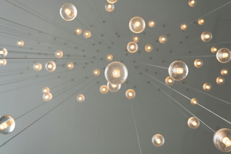 Hier ist ein Alt-Text für das Bild: "Moderne Licht-Installation an einer grauen Decke, von unten fotografiert. Zahlreiche transparente kugelförmige Leuchten in verschiedenen Größen hängen an dünnen transparenten Kabeln herab und erzeugen warmes, goldenes Licht. Die Perspektive verläuft nach oben zur Decke, wodurch eine eindrucksvolle Tiefenwirkung entsteht. Die klaren Kabel laufen von den Leuchten radiusartig zur Deckenfläche. Die Installation wirkt minimalistisch, kunstvoll und raumschaffend. Das warme Licht kontrastiert elegant mit der kühlen grauen Deckenoberfläche. Die Szene vermittelt moderne Raumgestaltung und künstlerisches Design.