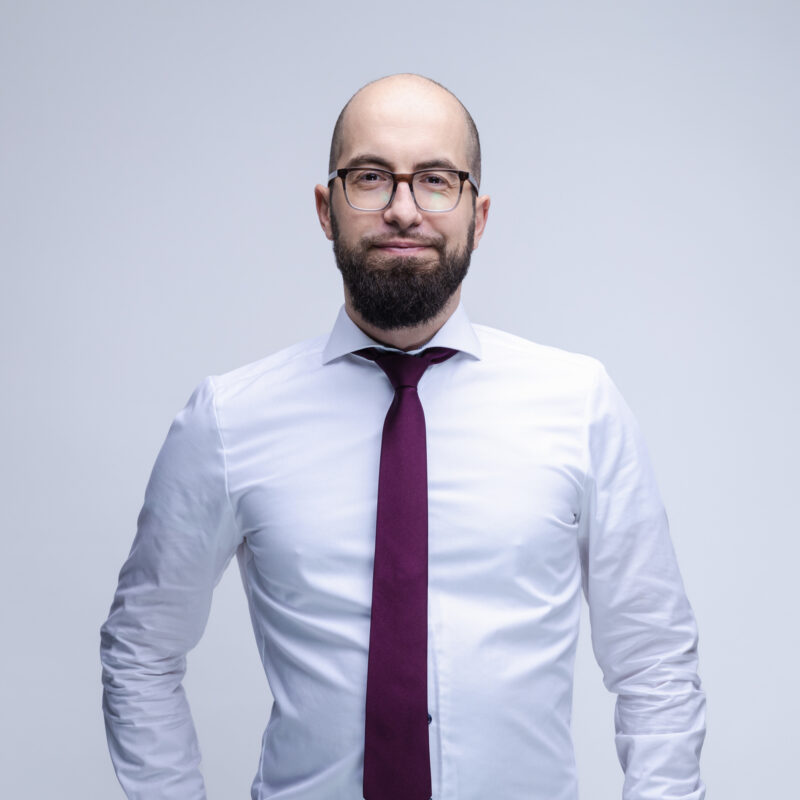 Porträtfoto von Gregor Theado mit Glatze, dunklem Bart und eckiger Brille vor hellgrauem Hintergrund. Er trägt ein weißes Hemd und eine dunkelrote Krawatte und blickt leicht lächelnd in die Kamera. Die Hände hat er hinter dem Rücken verschränkt. Foto: Thomas Quinten.
