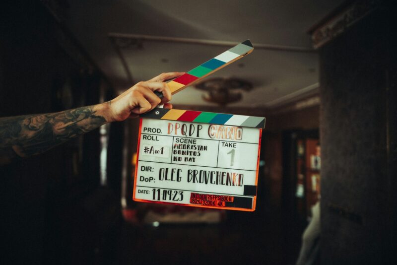 Eine tätowierte Hand hält eine Filmklappe in die Kamera. Auf der Klappe sind folgende Angaben zu lesen: Produktion „DPQDP CANO", Roll #A001, Szene „Amorestan Bonitos No Hay", Take 1, Regie und Kamera: Oleg Brovchenko, Datum: 11.09.23. Die Klappe hat einen bunten Streifen in Regenbogenfarben. Der Hintergrund ist dunkel und unscharf.