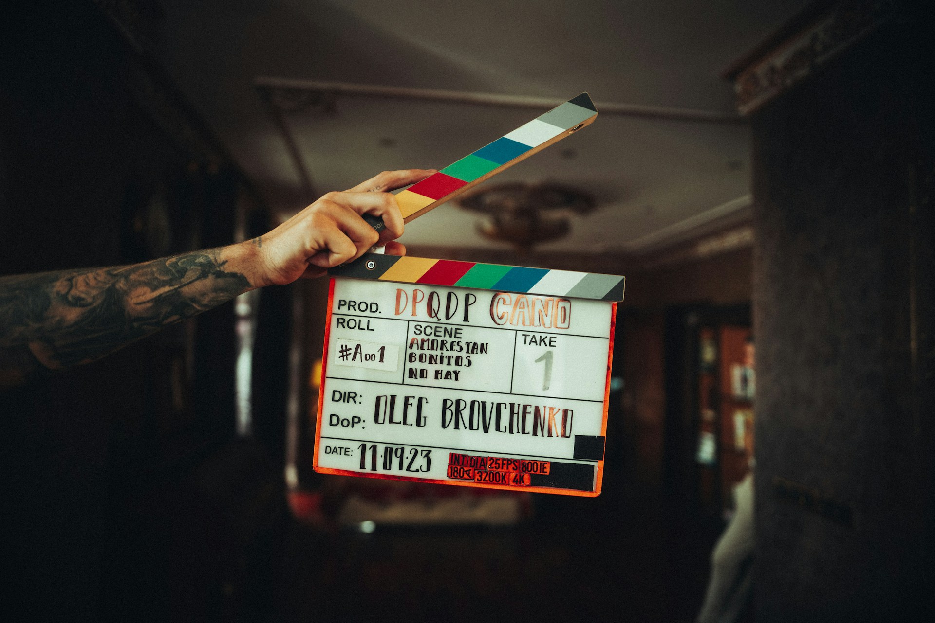 Eine tätowierte Hand hält eine Filmklappe in die Kamera. Auf der Klappe sind folgende Angaben zu lesen: Produktion „DPQDP CANO", Roll #A001, Szene „Amorestan Bonitos No Hay", Take 1, Regie und Kamera: Oleg Brovchenko, Datum: 11.09.23. Die Klappe hat einen bunten Streifen in Regenbogenfarben. Der Hintergrund ist dunkel und unscharf.