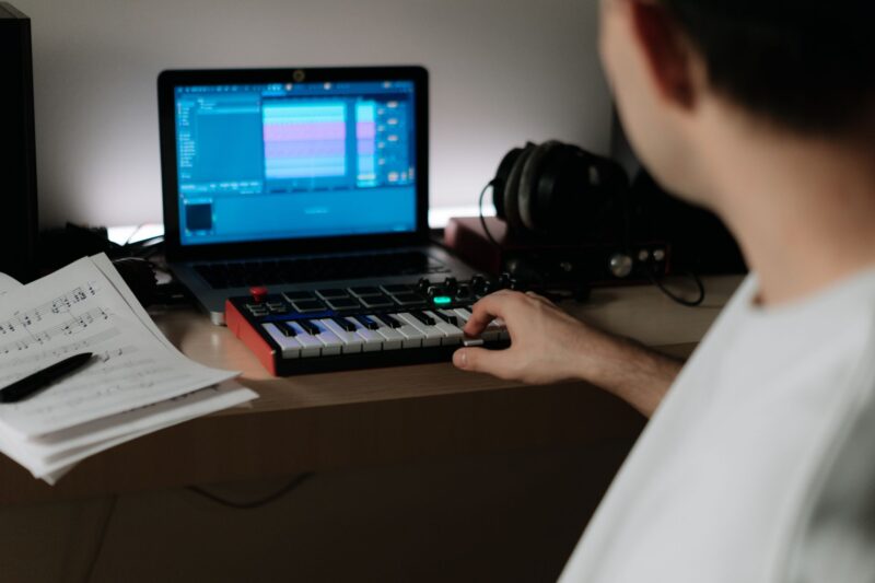 ine Person von hinten sitzt an einem Holzschreibtisch und bedient ein kompaktes MIDI-Keyboard mit schwarzem und rotem Gehäuse. Auf dem Laptop im Hintergrund ist eine Digital Audio Workstation (DAW) mit Spurspuren geöffnet. Rechts liegt ein schwarzes Over-Ear-Kopfhörerset, links befinden sich handgeschriebene Notenblätter und ein Stift. Das Bild zeigt eine typische Home-Studio-Produktionsatmosphäre.