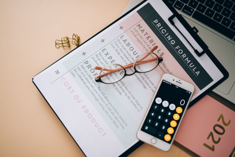 Draufsicht auf einen beigen Schreibtisch mit verschiedenen Arbeitsmaterialien. Im Mittelpunkt liegt ein aufgeschlagenes Notizbuch mit einem Arbeitsblatt zur „Pricing Formula", das Kategorien wie Materials, Labour, Expenses, Profit und Cost of Product enthält. Darauf liegen eine runde Brille und ein Smartphone mit geöffneter Taschenrechner-App. Daneben befinden sich goldene Büroklammern, ein rosa Notizbuch mit der Aufschrift „2021" sowie ein aufgeklappter Laptop. Das Bild vermittelt eine Arbeitsatmosphäre zum Thema Kalkulation und Preisgestaltung.