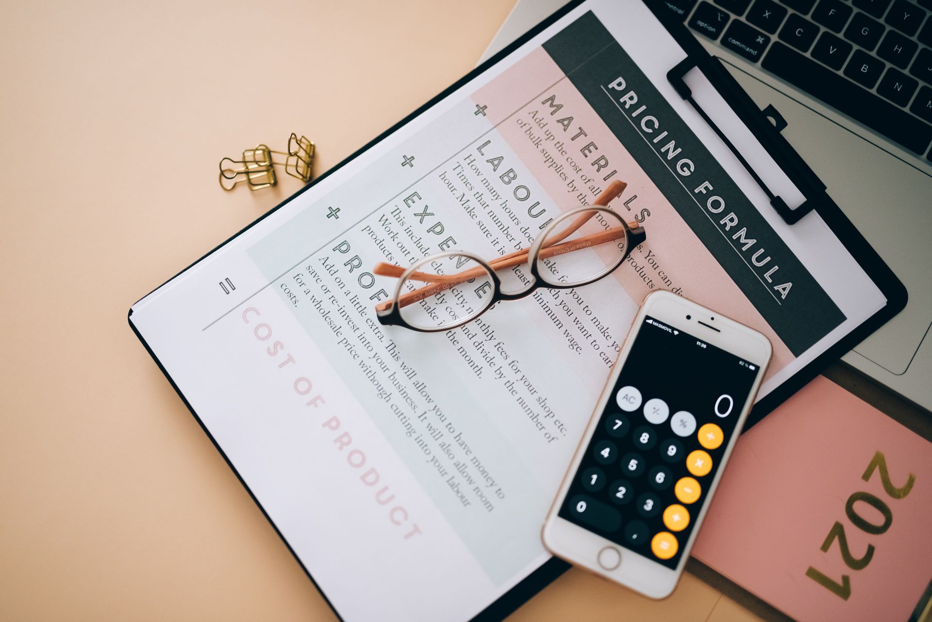 Draufsicht auf einen beigen Schreibtisch mit verschiedenen Arbeitsmaterialien. Im Mittelpunkt liegt ein aufgeschlagenes Notizbuch mit einem Arbeitsblatt zur „Pricing Formula", das Kategorien wie Materials, Labour, Expenses, Profit und Cost of Product enthält. Darauf liegen eine runde Brille und ein Smartphone mit geöffneter Taschenrechner-App. Daneben befinden sich goldene Büroklammern, ein rosa Notizbuch mit der Aufschrift „2021" sowie ein aufgeklappter Laptop. Das Bild vermittelt eine Arbeitsatmosphäre zum Thema Kalkulation und Preisgestaltung.