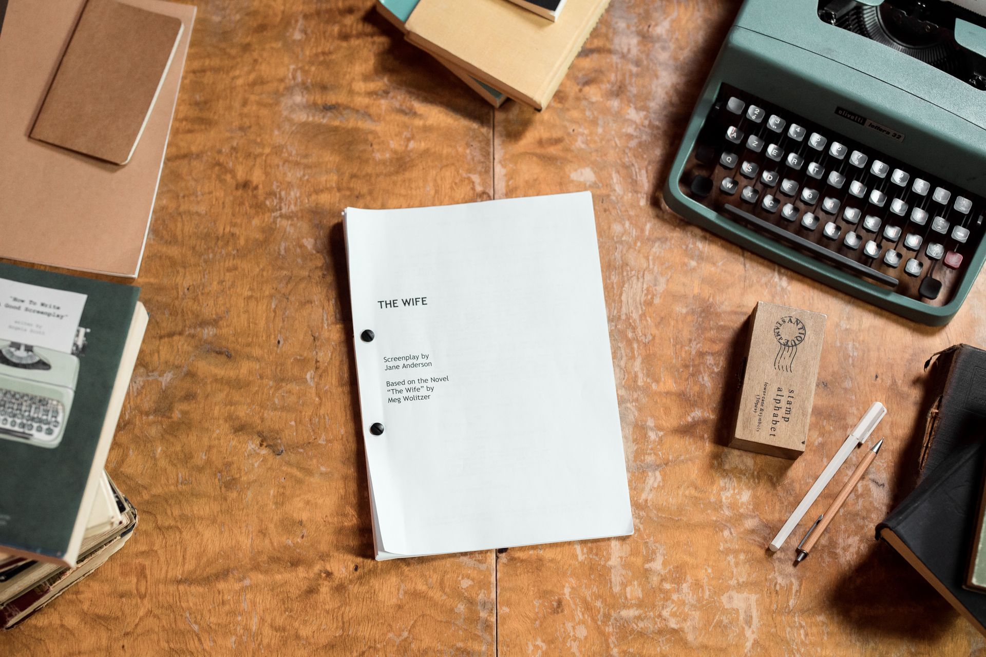 Alt-Text: Draufsicht auf einen Holzschreibtisch mit einem aufgeklappten Drehbuch im Mittelpunkt. Das Deckblatt trägt den Titel „THE WIFE – Screenplay by Jane Anderson, Based on the Novel 'The Wife' by Meg Wolitzer". Rechts oben steht eine türkisfarbene Olivetti-Schreibmaschine. Daneben liegen ein Holzstempel, ein Bleistift und ein Kugelschreiber sowie ein schwarzes Notizbuch. Links sind Bücher gestapelt, darunter eines mit dem Titel „How To Write Good Screenplay". Das Arrangement vermittelt eine nostalgische Schreibatmosphäre.