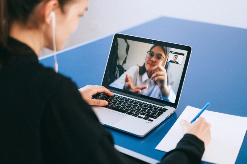 Eine Person von hinten sitzt an einem blauen Tisch vor einem Laptop und nimmt an einem Videoanruf teil. Sie trägt In-Ear-Kopfhörer und hält in der rechten Hand einen Stift über einen Block mit Notizen. Auf dem Laptopbildschirm ist eine junge Frau mit Brille und weißem Hemd zu sehen, die gestikulierend spricht. Das Bild vermittelt eine Online-Meeting- oder Mentoringatmosphäre.