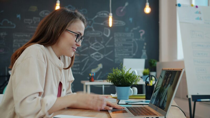 Eine junge Frau mit langen braunen Haaren und schwarzer Brille sitzt lächelnd vor einem Laptop und nimmt an einem Videogespräch teil. Sie trägt ein cremefarbenes Hemd und sitzt an einem Holztisch. Im Hintergrund ist eine schwarze Kreidetafel mit bunten Diagrammen und Notizen zu sehen, dazu eine kleine Pflanze, Hängeleuchten und ein Whiteboard. Die Atmosphäre wirkt kreativ und modern.