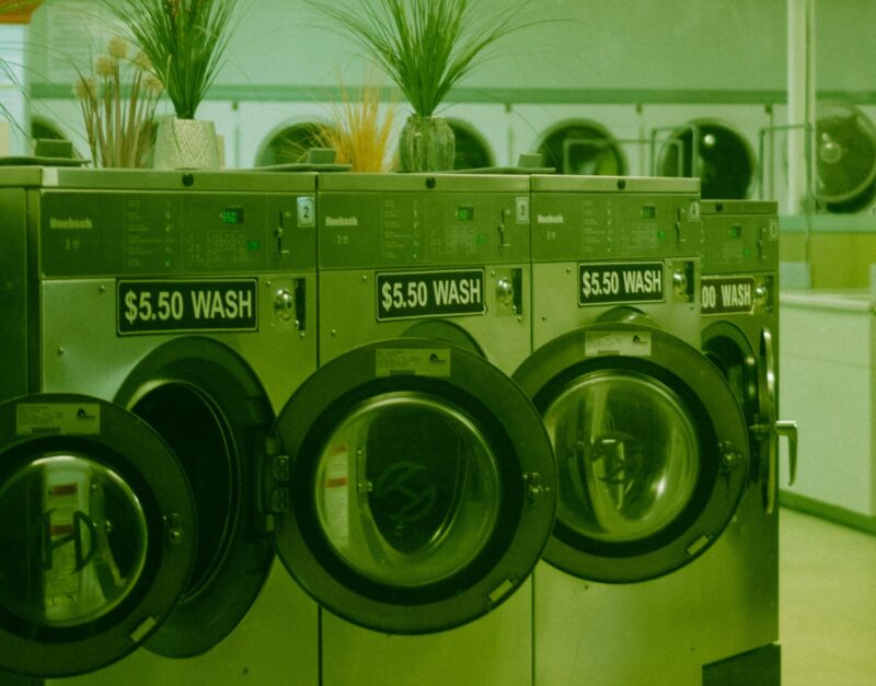 Innenansicht eines Waschsalons mit einer Reihe von Frontlader-Waschmaschinen der Marke Huebsch. Jede Maschine trägt ein schwarzes Schild mit der Aufschrift „$5.50 WASH". Auf den Geräten stehen Dekorationspflanzen in Vasen. Das gesamte Bild ist in einem markanten Grünstich gehalten, der an eine analoge Filmfotografie erinnert. Im Hintergrund sind weitere Maschinen und der helle Raum des Laundromats zu sehen.